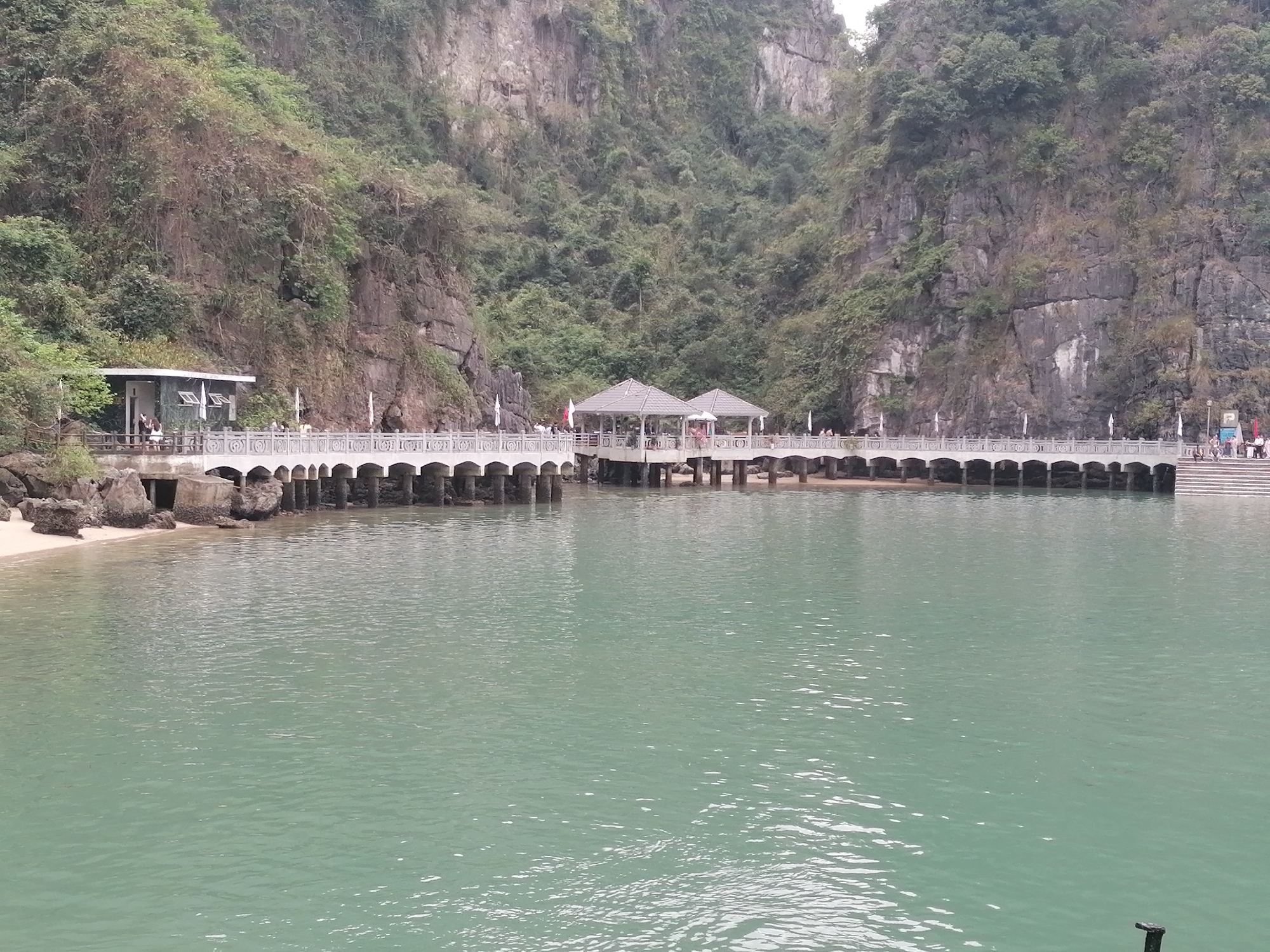 This wide-angle shot captures a serene coastal scene in Hồng Gai, Vietnam, characterized by dramatic limestone karsts and calm waters. In the foreground, a tranquil expanse of greenish-blue water reflects the diffused light.
Stretching across the midground is a long, curved pier or walkway, constructed with multiple arched supports. The pier features ornate white or light grey railings and serves as a public access point. On the far left of the pier, a modern-looking building with a dark roof and white walls sits on a small sandy beach, partially obscured by rocks and lush foliage. Further along the pier, two covered gazebos or shelters with light-colored roofs provide shade and gathering points for visitors. To the far right, the pier terminates in a wide set of stairs leading up to a higher platform.
Numerous people are visible along the pier, walking, standing, and congregating around the buildings and gazebos, suggesting a popular tourist destination. Most appear to be casually dressed, engaging in sightseeing or waiting activities.
The background is dominated by towering, steep limestone mountains, typical of the region, densely covered in vibrant green vegetation. The sky is not visible, with the majestic karsts filling the upper portion of the frame. The lighting suggests it is daytime, possibly an overcast day, given the soft, even illumination. A faint "P" symbol on a white sign is discernible on the right side of the pier.