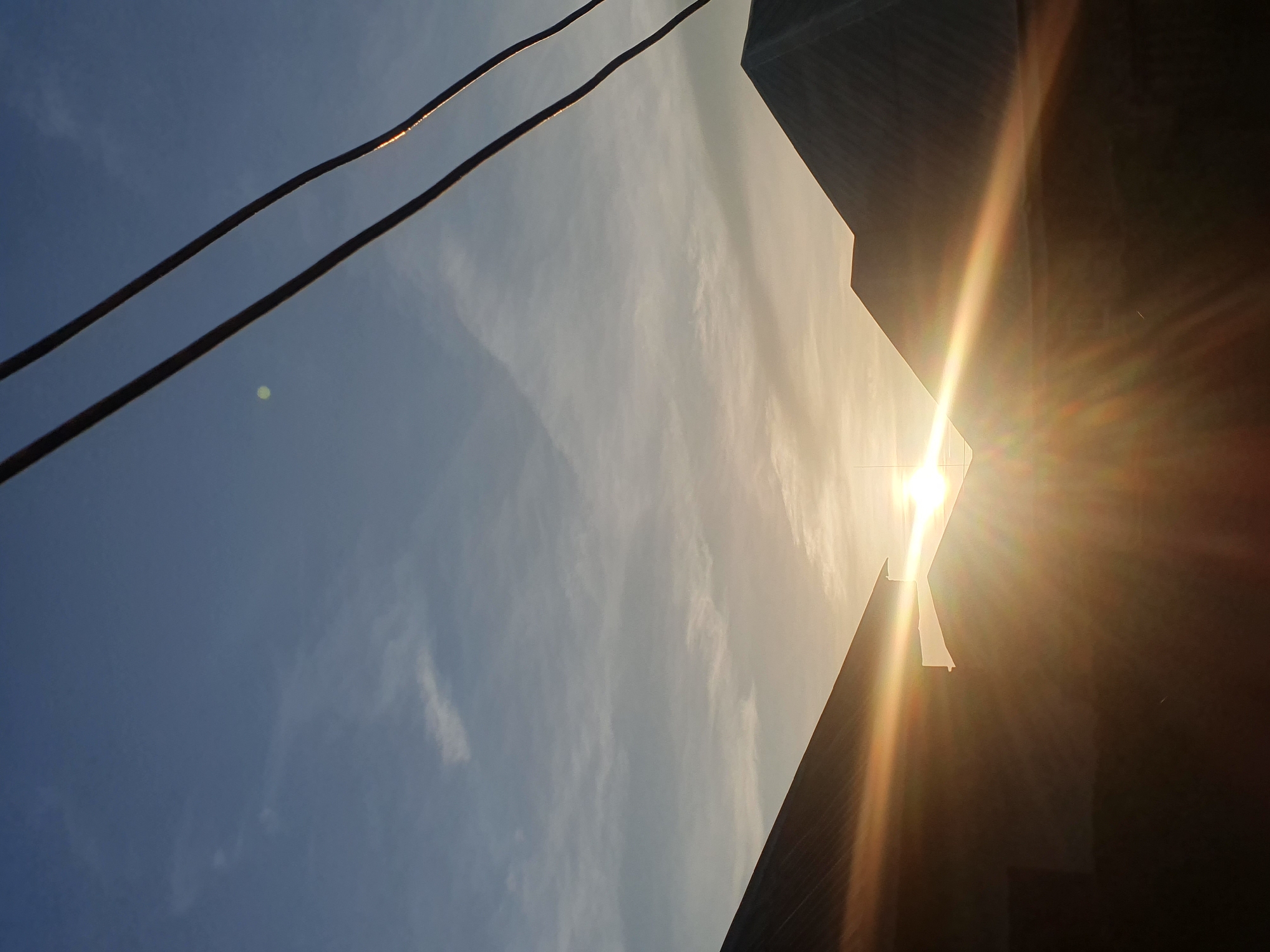The image depicts a sunset over rooftops in Gura Top, Nigeria. The sun is low on the horizon, casting a warm, golden light across the scene. Two silhouetted rooftops frame the sun, with the light flaring between them. In the foreground, two dark power lines stretch diagonally across the sky. The sky is a pale blue, with wispy white clouds. The overall impression is one of a serene evening in a residential area.