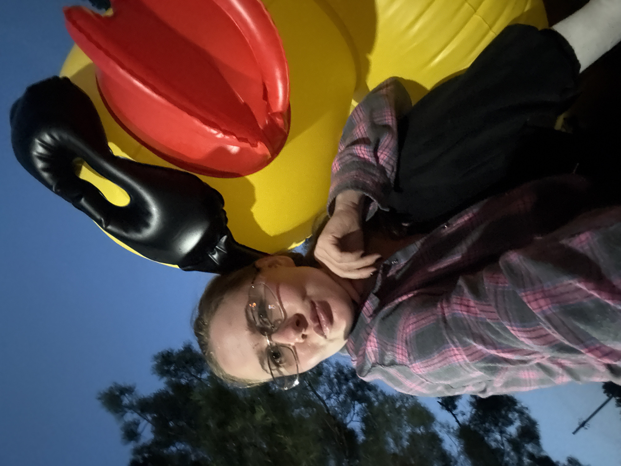A woman with blonde hair and glasses is lying on an inflatable duck. She is wearing a pink and gray plaid shirt and black pants. The inflatable duck is yellow with a red beak and black eyes. The background is a dark blue sky with some trees. The image appears to have been taken at night. The location is the city of Chaykovsky, Russia.