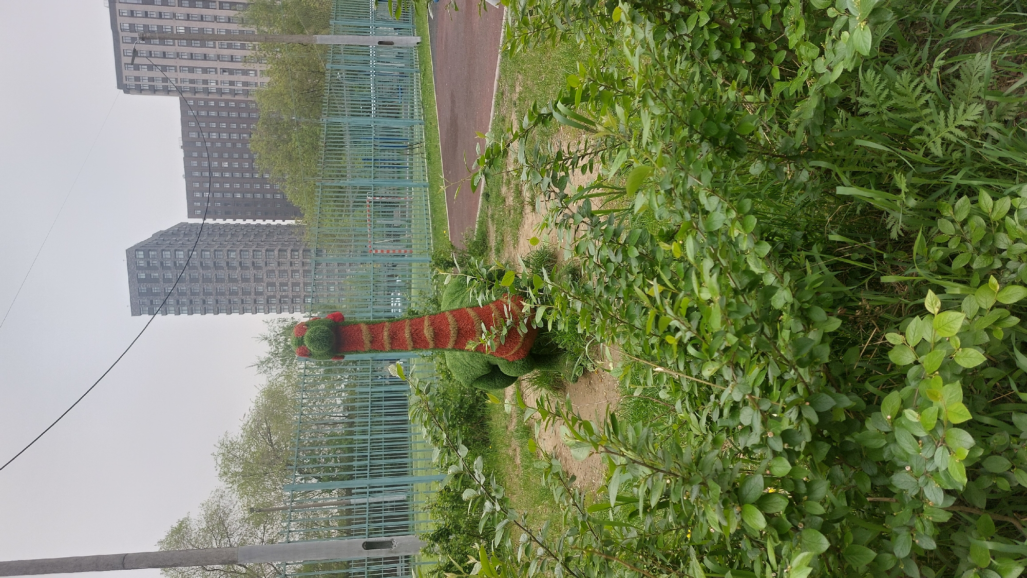 The image shows a park scene in Moscow, Russia. A topiary shaped like a dinosaur sits near a green fence. Behind the fence, a red path runs alongside it. Tall apartment buildings are visible in the background under a cloudy sky. The topiary has a green rounded base with a long neck striped in red and brown. Lush green bushes and trees surround the dinosaur sculpture, partially obscuring it. An overhead power line is visible in the upper left corner. The scene appears to be in a well-maintained urban park.