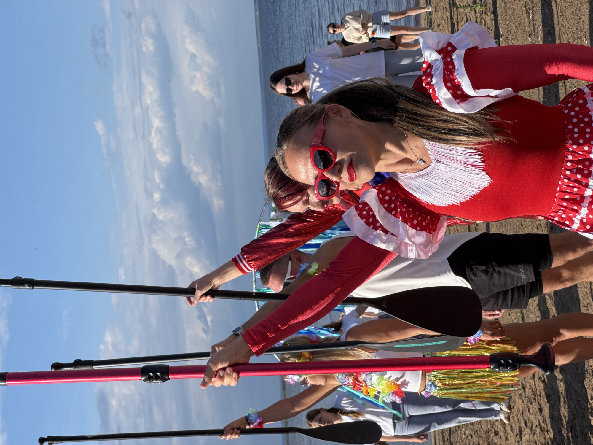 The image depicts a group of people on a sandy shore near a body of water, likely a river or lake, under a partly cloudy sky. The location appears to be the Kama River embankment in Chaikovsky, Russia, given the distinctive landscape and the context of the event.
In the foreground, a woman with blonde hair pulled into a ponytail wears red-rimmed sunglasses and a festive red and white polka dot dress with ruffled sleeves and fringe detailing around the neckline. She is holding a black paddle, suggesting participation in a water sport like paddleboarding.
To her left, another woman, partially visible, wears a similar red, ruffled garment and a festive lei around her neck. Behind her, a young girl with blonde hair in braids also holds a paddle.
Further back, several other individuals are visible, some holding paddles. One man is wearing white sunglasses and a white t-shirt. A woman in the background wears a light-colored hoodie and denim shorts.
The atmosphere suggests a celebratory event or a group activity, possibly related to a festival or sports competition, taking place on a sunny day. The paddles are prominent objects, indicating a shared activity. The backdrop consists of the calm water of the Kama River and a blue sky with scattered clouds.