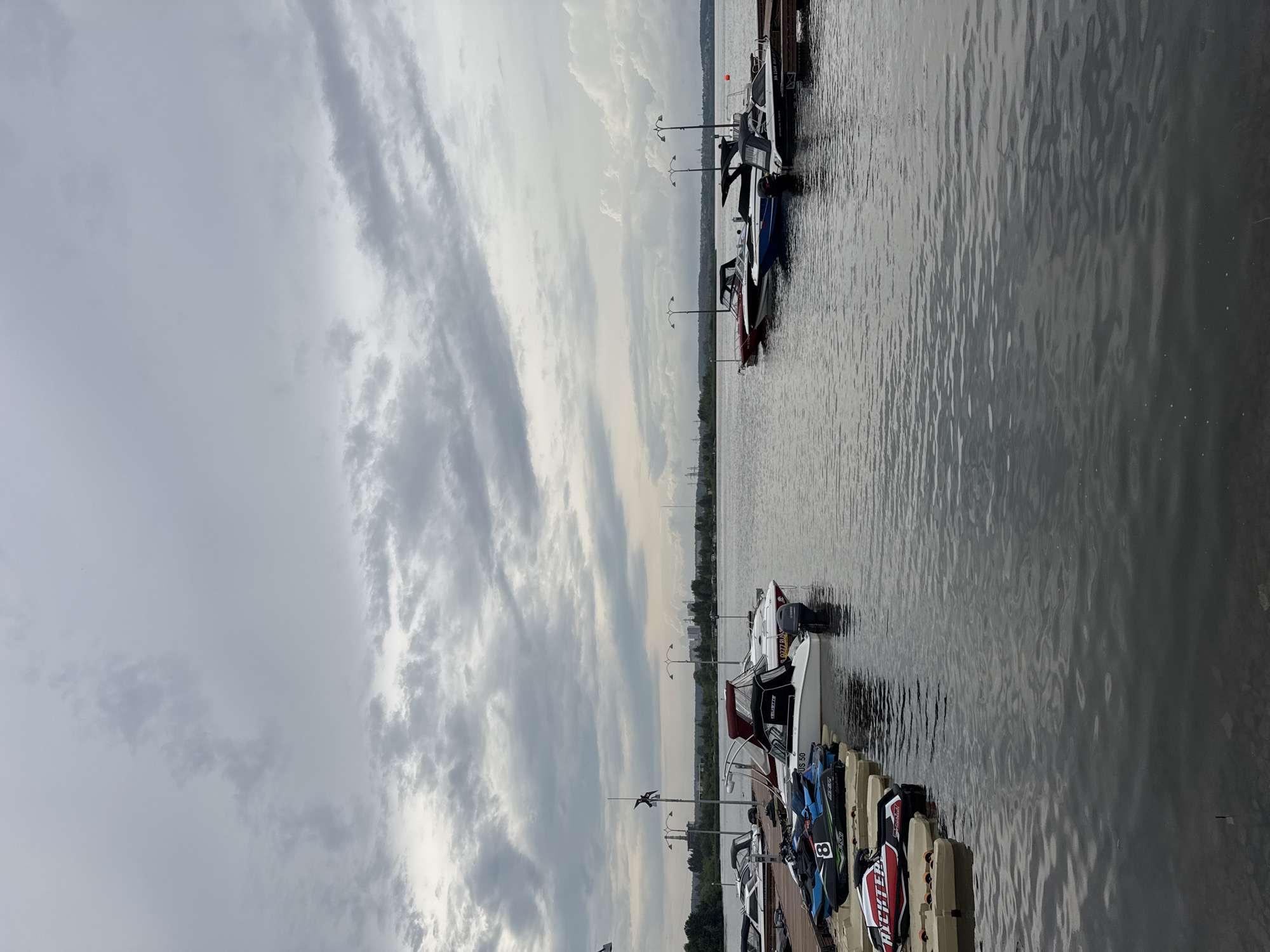 The image depicts a waterfront scene at a marina, likely in Izhevsk, Russia, given the context. The sky is overcast with dramatic, textured clouds. A calm body of water fills the lower half of the frame, with gentle ripples on its surface. Several boats are docked along a wooden pier in the foreground and mid-ground.
On the left side of the pier, there are several jet skis, one of which is black and blue with the number "8" prominently displayed in white. Adjacent to the jet skis are smaller recreational boats, some with covered canopies. Further down the pier, a larger white motorboat with a red stripe and a dark canopy is docked.
On the right side of the pier, more motorboats are moored. These boats vary in size and color, including a red boat and several white boats with dark accents. Streetlights on poles are visible along the pier and in the background.
The background shows a tree-lined shore with industrial structures and smokestacks in the distance, suggesting a mix of natural and urban elements in the vicinity. The overall atmosphere is subdued due to the cloudy weather. There are no people visible in the image.