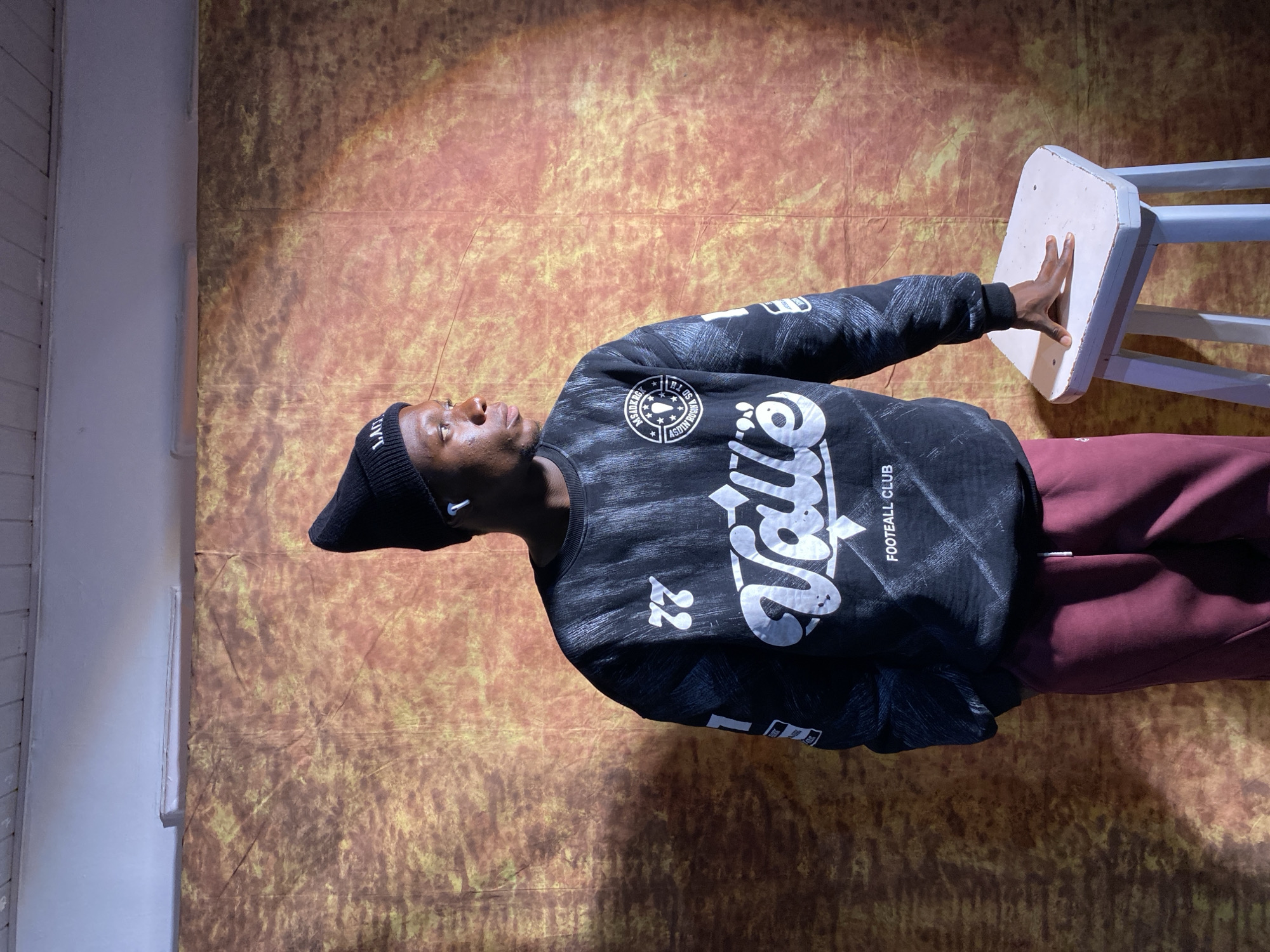 The image depicts a young Black male standing in what appears to be an indoor studio setting. He is wearing a black beanie, a black sweatshirt with white graphics that include the word "Valle" and "FOOTBALL CLUB", and maroon sweatpants. He has white earbuds in his ears. He is looking to his right, with his right hand resting on a small, white stool. The background is a textured fabric in shades of brown and yellow, with a spotlight creating a dramatic effect. The ceiling above is white with a fluorescent light fixture. There is no indication of the specific location in the city of Barakin Akawo, Nigeria.