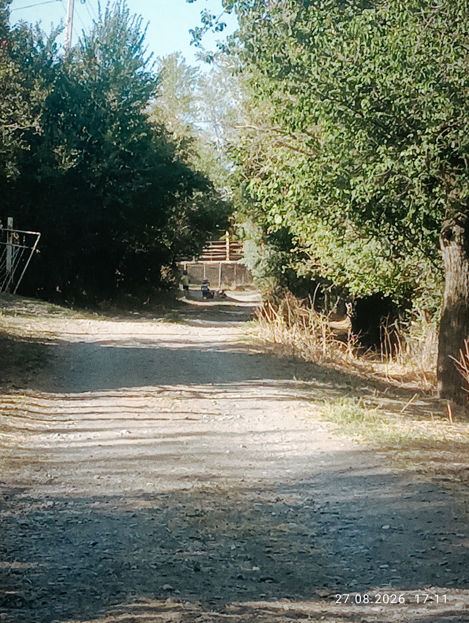 The image depicts a dirt path winding through a wooded area. On the left side of the path, there's a metal fence. Further up the path, a few individuals are visible, and some appear to be near a structure with a fence. The trees are lush and green, suggesting a warm season. The path itself is uneven and appears to be made of gravel and dirt. Sunlight filters through the trees, creating shadows on the ground. The overall scene conveys a sense of an outdoor, possibly rural or park-like setting. The timestamp in the lower right corner indicates the photo was taken on August 27, 2026, at 17:11. It is not possible to determine the specific location within Ak-Took, Kyrgyzstan from the visual information provided.