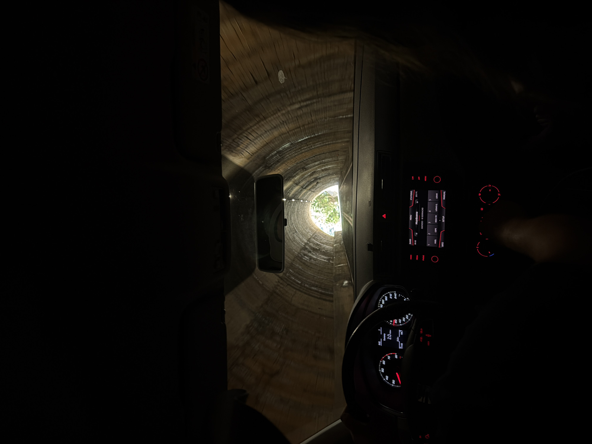 Captured from inside a vehicle, the image presents a view through a tunnel, possibly constructed from wood or stone, terminating with a patch of light indicative of an exit surrounded by foliage. The tunnel's structure is circular or arched, appearing to be weathered or aged. The vehicle's dashboard is visible, featuring a steering wheel and an electronic display panel with illuminated controls and information. A smartphone is attached to the dashboard or windshield. The environment is dark inside the vehicle, contrasted by the light source at the end of the tunnel. Given the information available, the location might be in Escorca, Spain, but direct evidence of this is not visible in the image. There are no visible people.