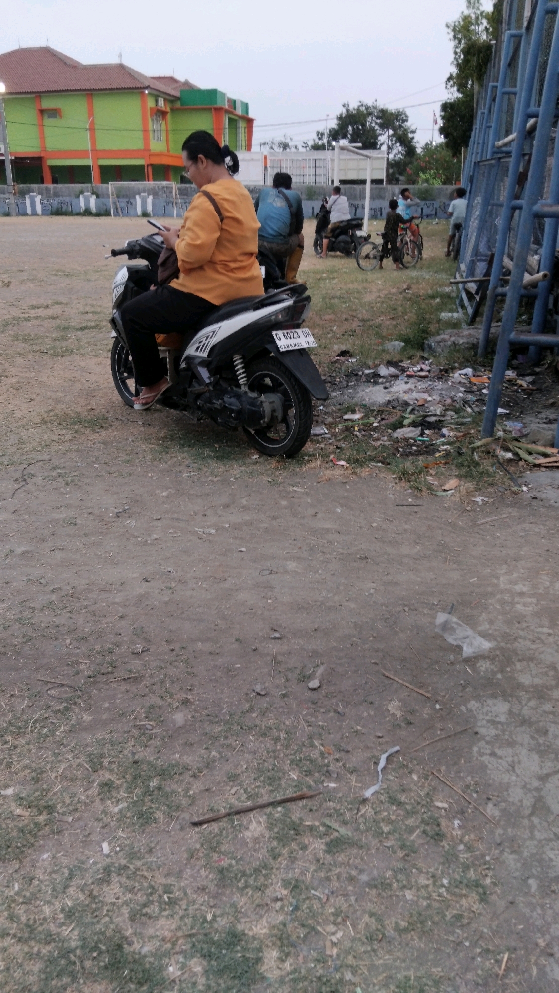 An exterior, low-angle, full shot shows a woman on a scooter in what appears to be a public space in Tegal, Indonesia. The woman is wearing an orange shirt and black pants and is looking at her phone. The scooter is black and white and has a license plate that reads "G 6023 GH". In the background, there is a building with a green and orange facade, and several people are visible on motorbikes and bicycles. There is also a blue metal structure in the right side of the frame. The ground is covered with dirt and dry grass, and there is some litter scattered around. The sky is overcast.