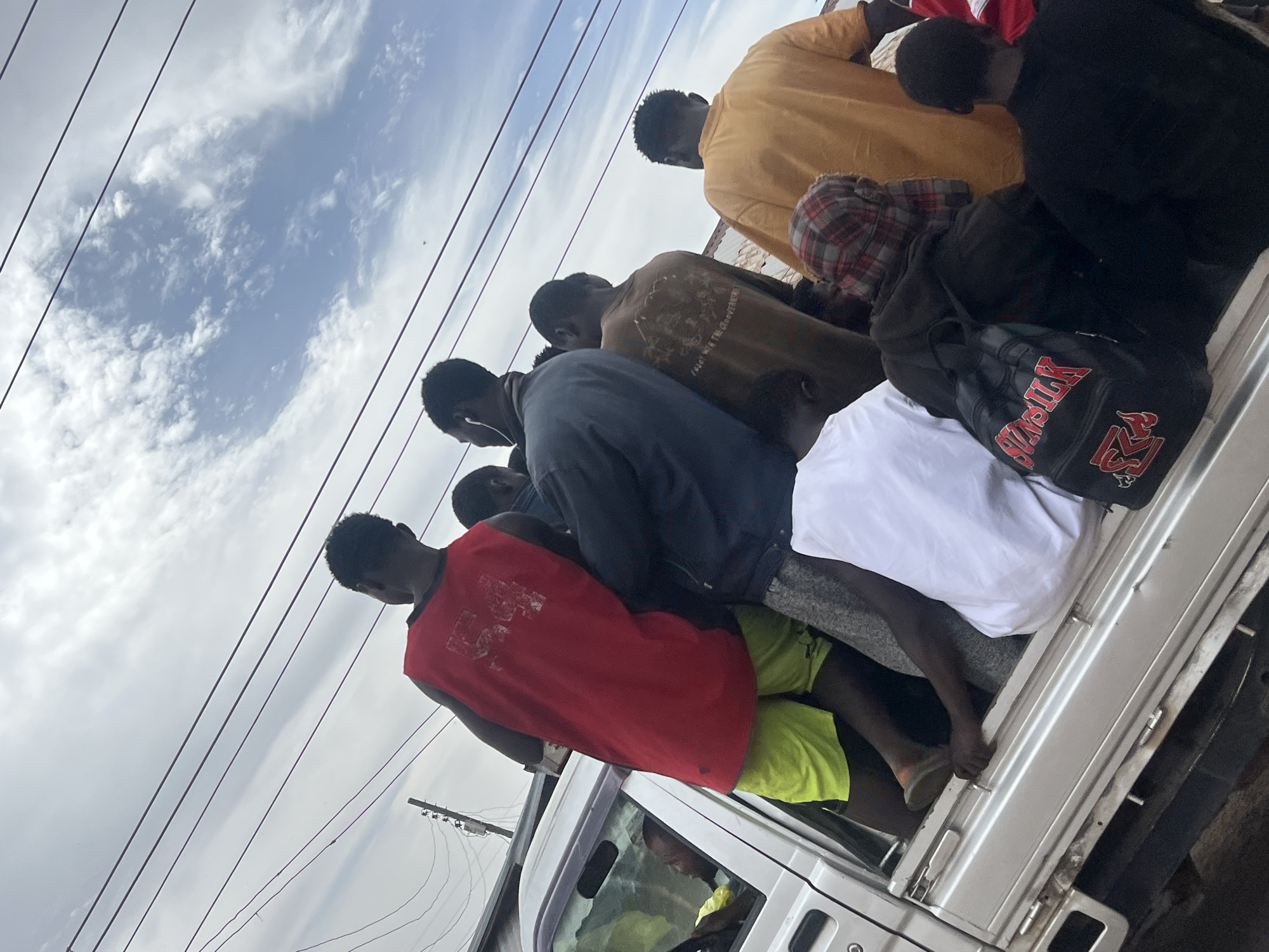 The image, captured with a tilted perspective, depicts approximately 10-12 young men or boys crowded into the open bed of a light-colored utility vehicle, possibly a pickup truck, under a bright, cloudy sky in Jos, Nigeria. The scene suggests communal or informal transportation.
The individuals are dressed casually in various t-shirts and tank tops (red, yellow, brown, dark blue, white), shorts (including neon yellow), and some long pants. One person wears a plaid shirt. They are positioned closely together, some standing, others crouching or sitting, facing generally towards the front of the vehicle. One person on the left side of the frame has white earbuds. The driver of the vehicle is partially visible through the front windshield.
The sky above is overcast with white cumulus clouds, revealing patches of blue. Multiple dark power lines are strung across the upper portion of the frame. Among the belongings is a black duffel bag with prominent red lettering, which appears to spell "SWASIKK" and includes an "R" or "M" logo. Partially visible text on a brown t-shirt reads "GOD IS THE GOVERN". The time of day is clearly daytime, and the weather is bright with cloud cover.