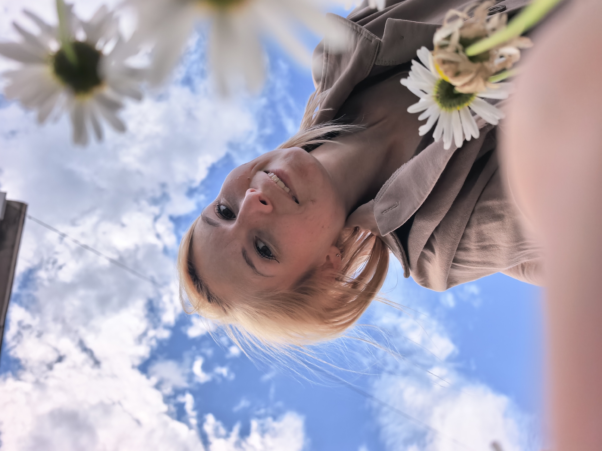 A low-angle, close-up, Dutch-angle selfie shows a young blonde woman looking up and smiling. She is wearing a light brown jacket and holding a white daisy. Two other daisies are visible in the foreground, out of focus. The background is a bright blue sky with fluffy white clouds. The image is oriented upside down, with the woman's head at the bottom of the frame and the sky at the top. The setting is an outdoor environment, possibly in a park or garden, in the city of Sterlitamak, Russia.