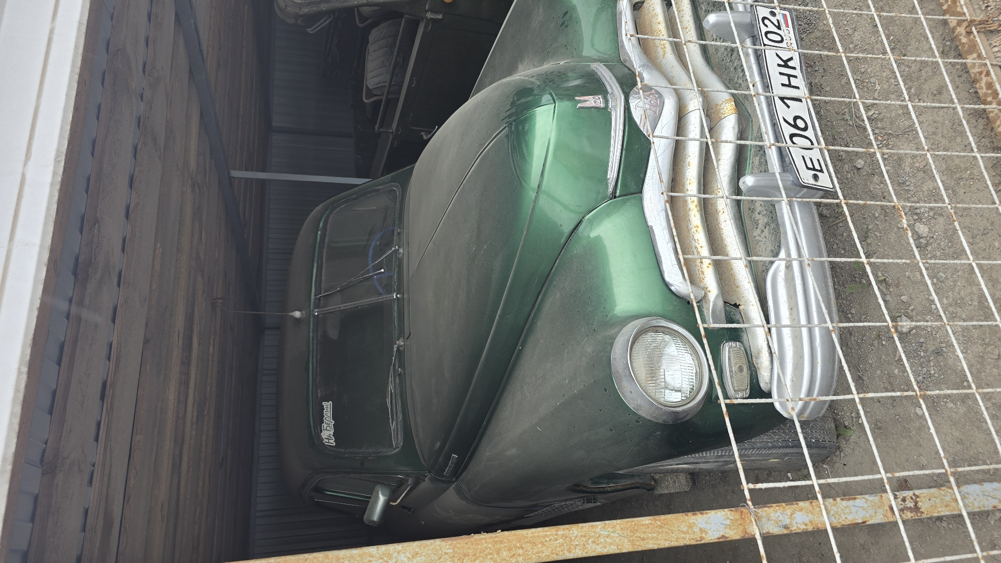 A green vintage truck is parked in a wooden structure, possibly a garage or barn. The truck is facing towards the right of the frame, with its front grill and headlights clearly visible. The grill is chrome and shows some signs of rust. A license plate with the text "E 061 HK 02" and "RUS" is attached to the front of the truck. The truck's hood is dusty, and the windshield is visible. There is a wire fence in the foreground, partially obscuring the view of the truck. On the side of the truck, near the driver's side door, a sticker with text that appears to be "НА БЕРЕГА" is visible. The setting appears to be outdoors in Bol'shoi Mashlyak, Russia, with a gravelly ground and some vegetation visible behind the fence. The lighting suggests it is daytime. No people are visible in the image.