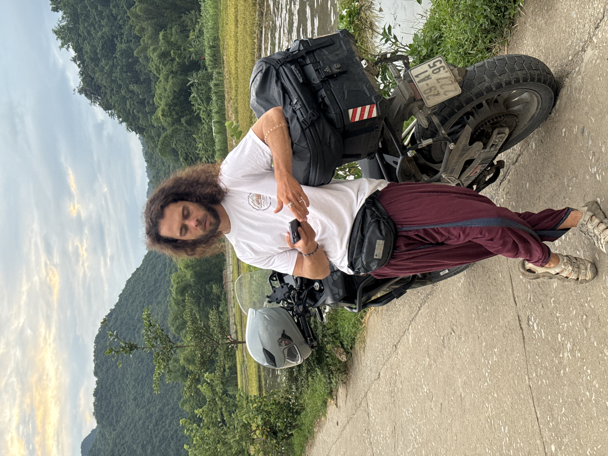 The image shows a young man with long, curly, dark hair and a beard, standing next to a motorcycle. He is wearing a white t-shirt with a circular graphic on it, maroon pants, and tan sandals. He has a black fanny pack around his waist and a black backpack on the back of the motorcycle. He is holding a black smartphone in his right hand. The motorcycle has a grey helmet on the handlebars.
The scene appears to be rural, with lush green hills in the background and a body of water or flooded rice paddies to the left. The sky is cloudy with some streaks of orange light, suggesting it might be sunrise or sunset. The ground is a paved road with some cracks. The motorcycle has a license plate that reads "56-G1 222.95". The overall impression is of a traveler taking a break during a journey in a scenic, natural setting. The man's posture and the relaxed way he is holding his phone suggest a moment of pause and perhaps contemplation.