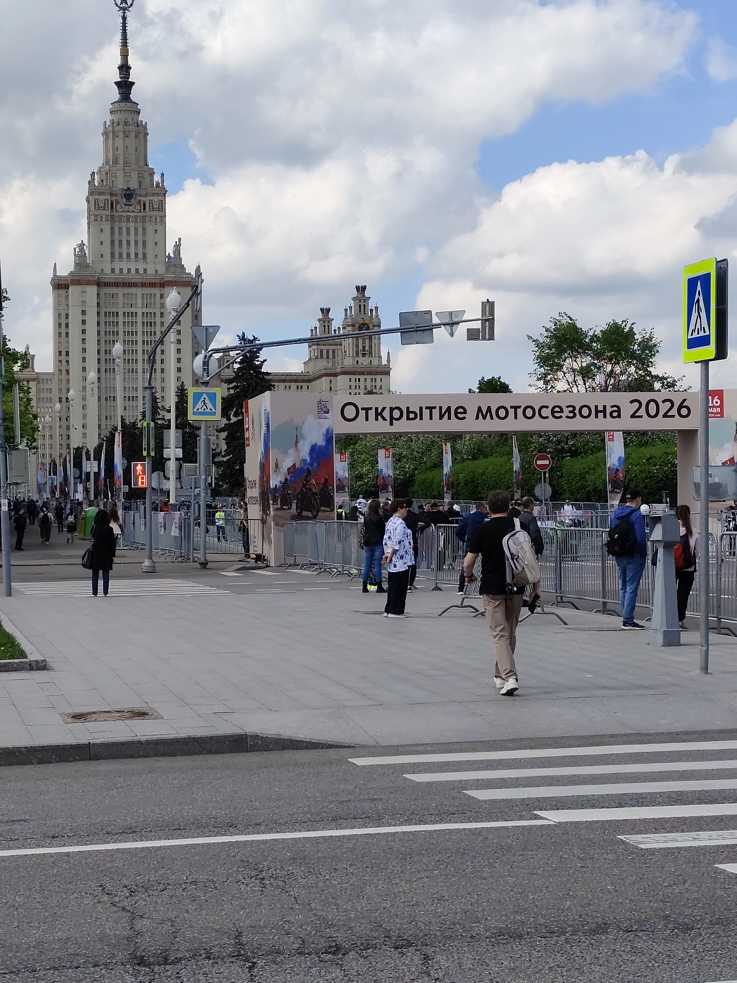 The image shows an urban scene in Moscow, Russia, featuring the iconic Moscow State University building in the background. In the foreground, a crosswalk spans a street with cars visible in the distance. A crowd of people stands on a wide sidewalk behind metal barriers. A banner stretched across the pedestrian path reads "Открытие мотосезона 2026" (Opening of the Motorcycle Season 2026). Several street signs, including pedestrian crossing signs and a red stop sign, are placed around the scene. The sky is partly cloudy. The photo appears to be taken during a public event or festival.