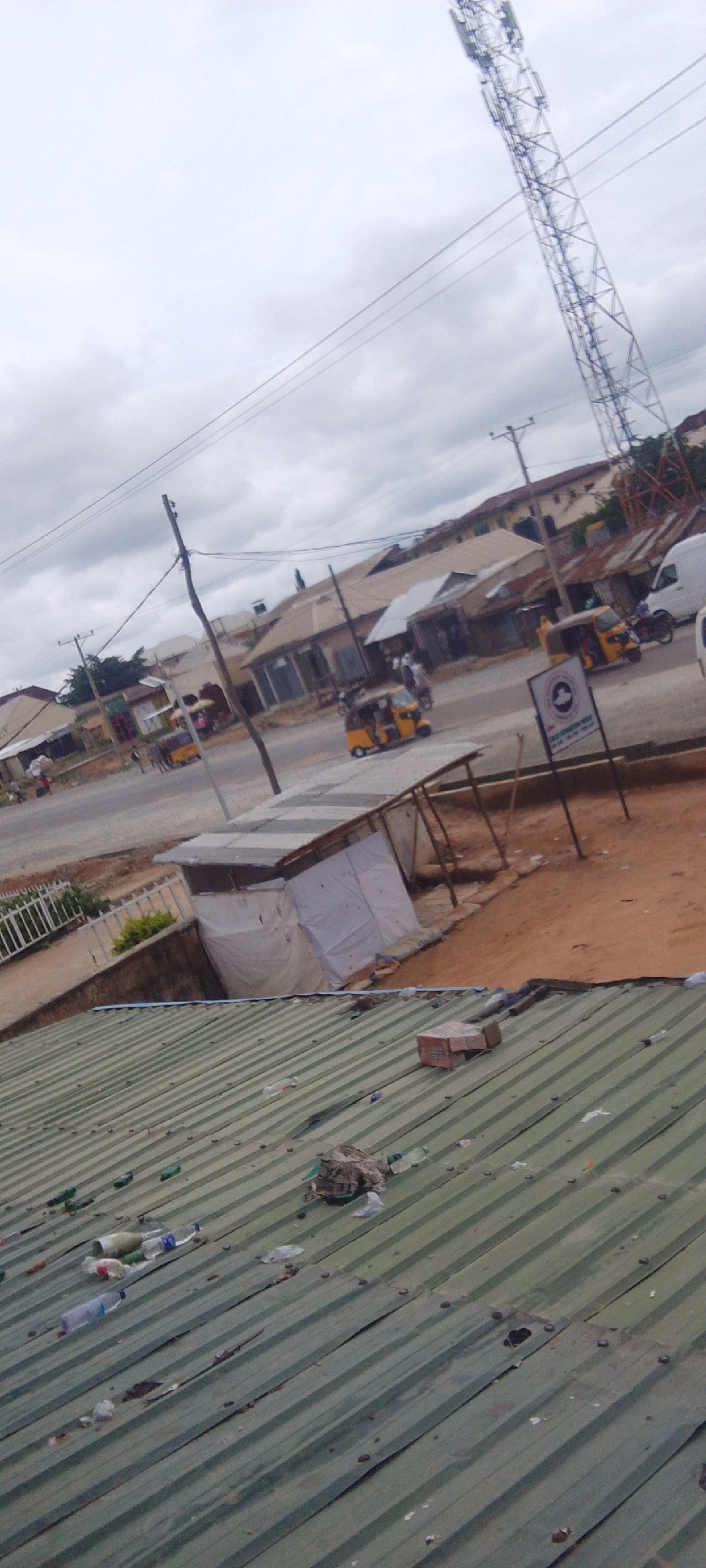 This is a medium-angle, daytime shot of a street scene in Suleja, Nigeria. The sky is overcast, suggesting cloudy weather. The foreground is dominated by a green corrugated metal roof littered with trash, including plastic bottles and what appears to be a cardboard box.
Beyond the roof, a dirt area with some sparse vegetation leads to a street. On the street, several three-wheeled motorized vehicles, commonly known as "Keke Napep" or "Marwa," are visible, some appearing to be in motion. There are also people walking and standing on the street. Buildings line the street, appearing to be a mix of shops and residences with corrugated metal roofs. In the background, a tall cell tower stands against the cloudy sky. A sign with circular emblem and text in English is visible on the right side of the street. The overall impression is that of a busy, somewhat unkempt urban environment during the day.