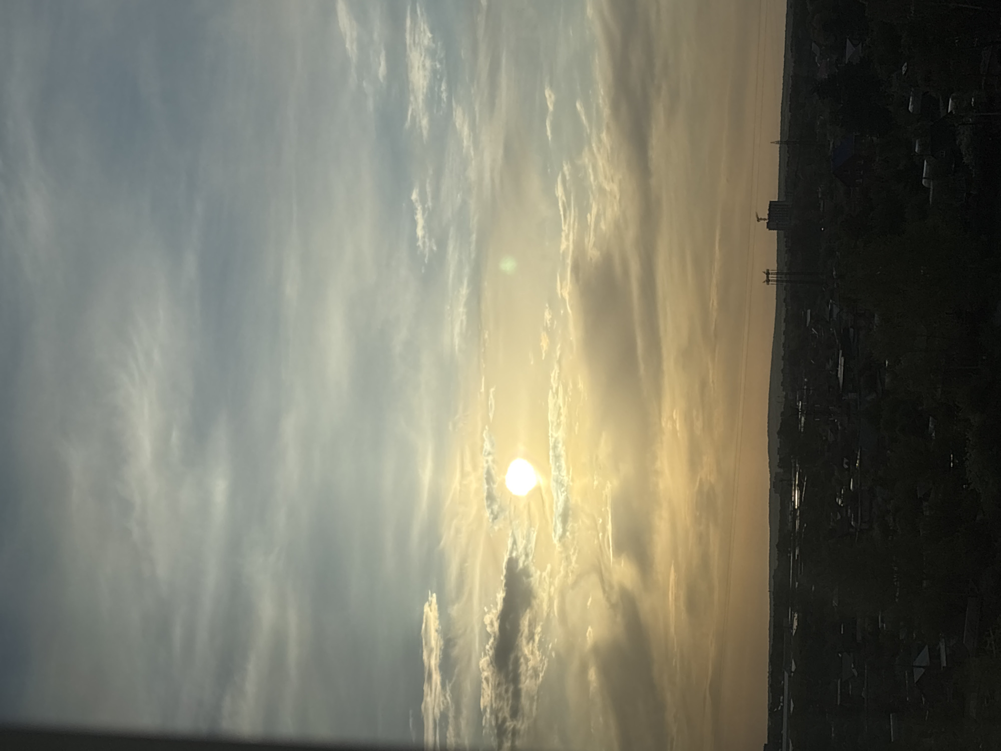 The image shows a sunset over Chaikovsky, Russia. In the foreground, there is a cityscape silhouette against the evening sky. The sun is visible, partially obscured by clouds. The sky has layered clouds with a gradient from lighter blue to the left to more golden tones towards the right where the sun is setting. Some industrial structures, possibly cranes, are visible in the distant cityscape. The image is taken from an elevated position.