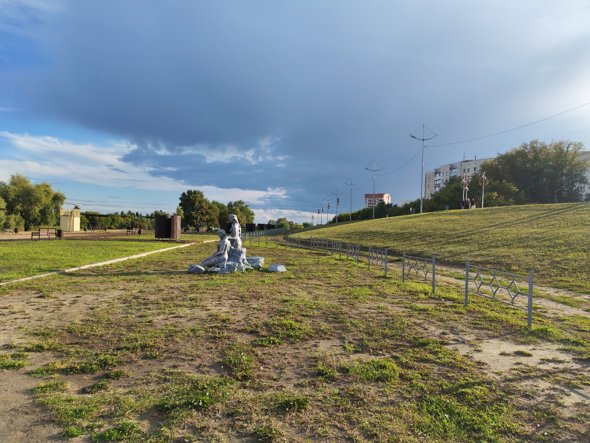 The image depicts a park-like area with a cloudy sky overhead. In the foreground, there is a sculpture of a seated figure with rocks at its base. To the left, a gravel path leads towards benches and a wooden structure. A metal fence runs along a grassy slope in the middle ground, separating a path from the lawn. In the background, buildings and trees are visible, along with streetlights. The scene appears to be in a public space, likely a park or recreational area. The sculpture is situated on a patch of dirt and grass. The overall ambiance suggests an outdoor setting in an urban or suburban environment.
