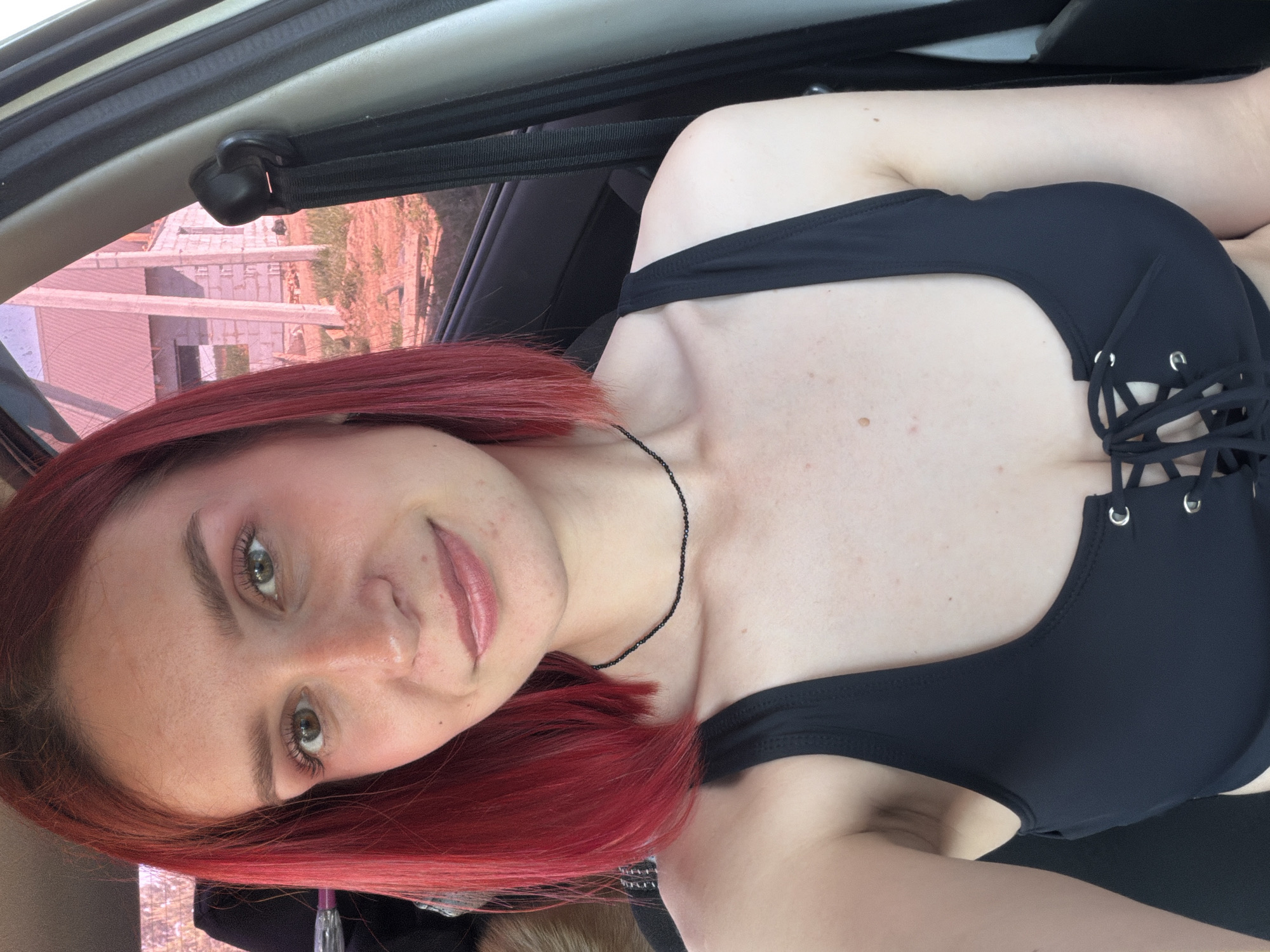 A selfie of a woman with red hair, wearing a black top, taken inside a car. The woman is looking at the camera with a slight smile. Outside the car window, there is a building under construction. Based on the angle and presence of a car interior, the subject is in Chaikovsky, Russia.