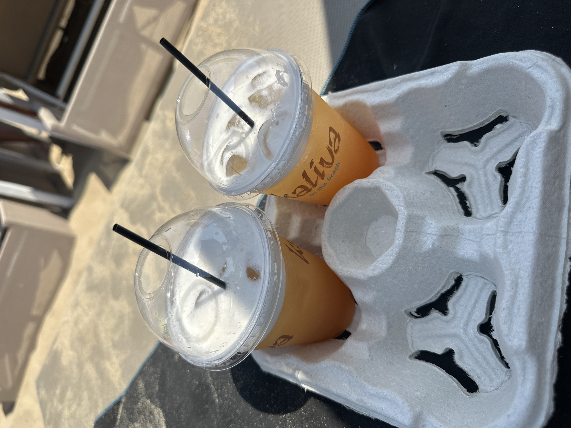 Two clear plastic cups with dome lids and black straws are filled with an orange beverage and topped with foam. The cups are placed in a white paper beverage carrier. The cups are labeled with "Maliva on the beach." The carrier and cups are positioned on a dark surface, possibly a towel, with a sandy or concrete-like background. In the blurred background, light-colored lounge chairs are visible. The lighting suggests an outdoor, daytime setting, likely a beach or poolside area in Ayia Napa, Cyprus.