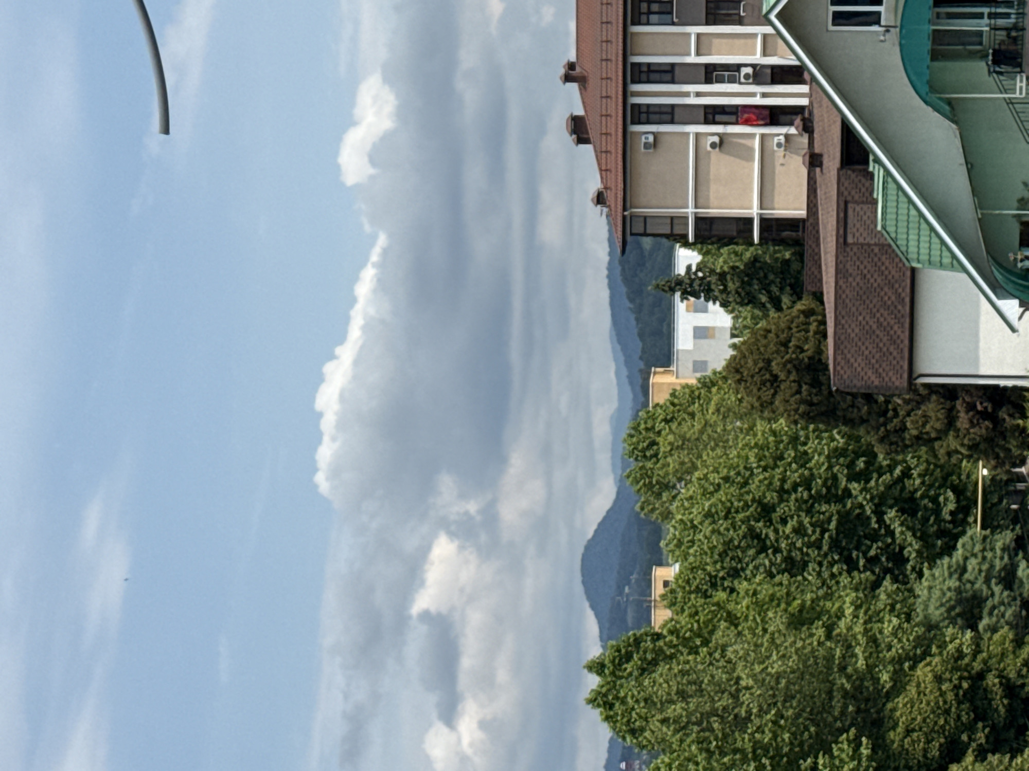 This is a daytime, eye-level, outdoor, medium shot of a cityscape in Sochi, Russia. In the foreground, a light gray curved structure, possibly a part of a lamp post or a cable, juts in from the right side of the frame. Behind it, a bright blue sky with scattered white clouds dominates the upper portion of the image. Below the sky, a range of green, tree-covered mountains forms the backdrop. In the mid-ground and foreground, several multi-story buildings with light-colored facades and brown-tiled roofs are visible. Trees with dense green foliage are scattered throughout the scene, obscuring some of the buildings. One building features a red flag or cloth hanging from a balcony. The scene appears to be part of a residential or commercial area.