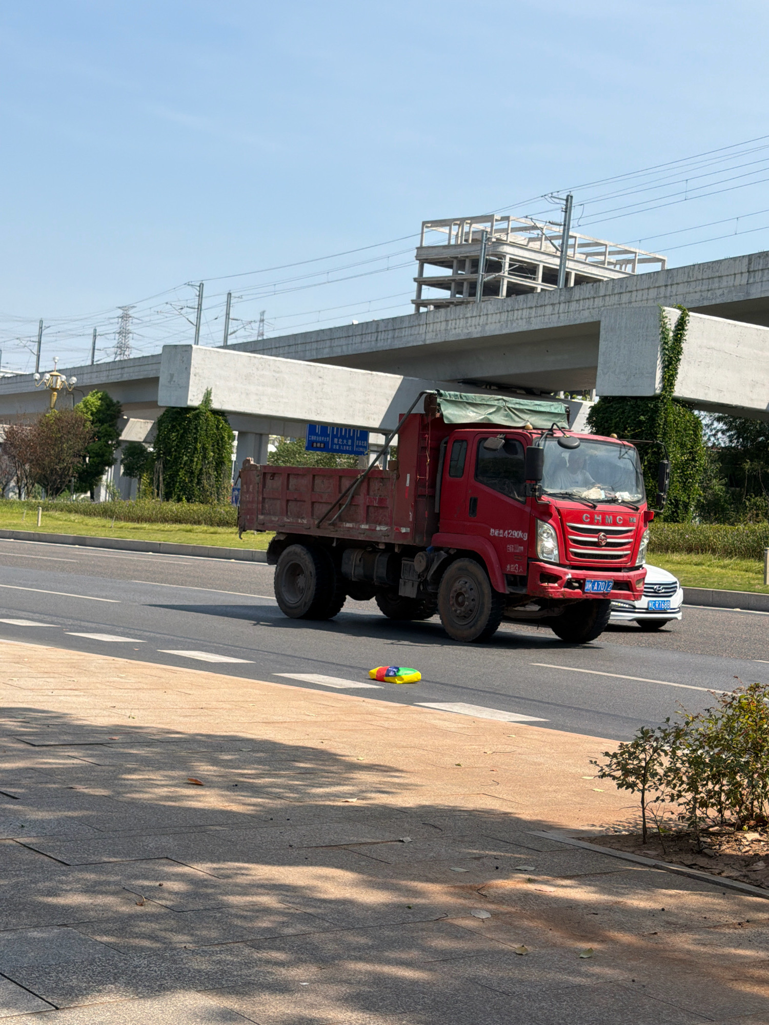 A red dump truck with a green tarp is driving on a road in Jiangjin, China. A white car is following behind the truck. In the foreground, a colorful children's toy is on the side of the road. In the background, there is an elevated highway under construction, with power lines and trees visible. The sky is clear and blue.