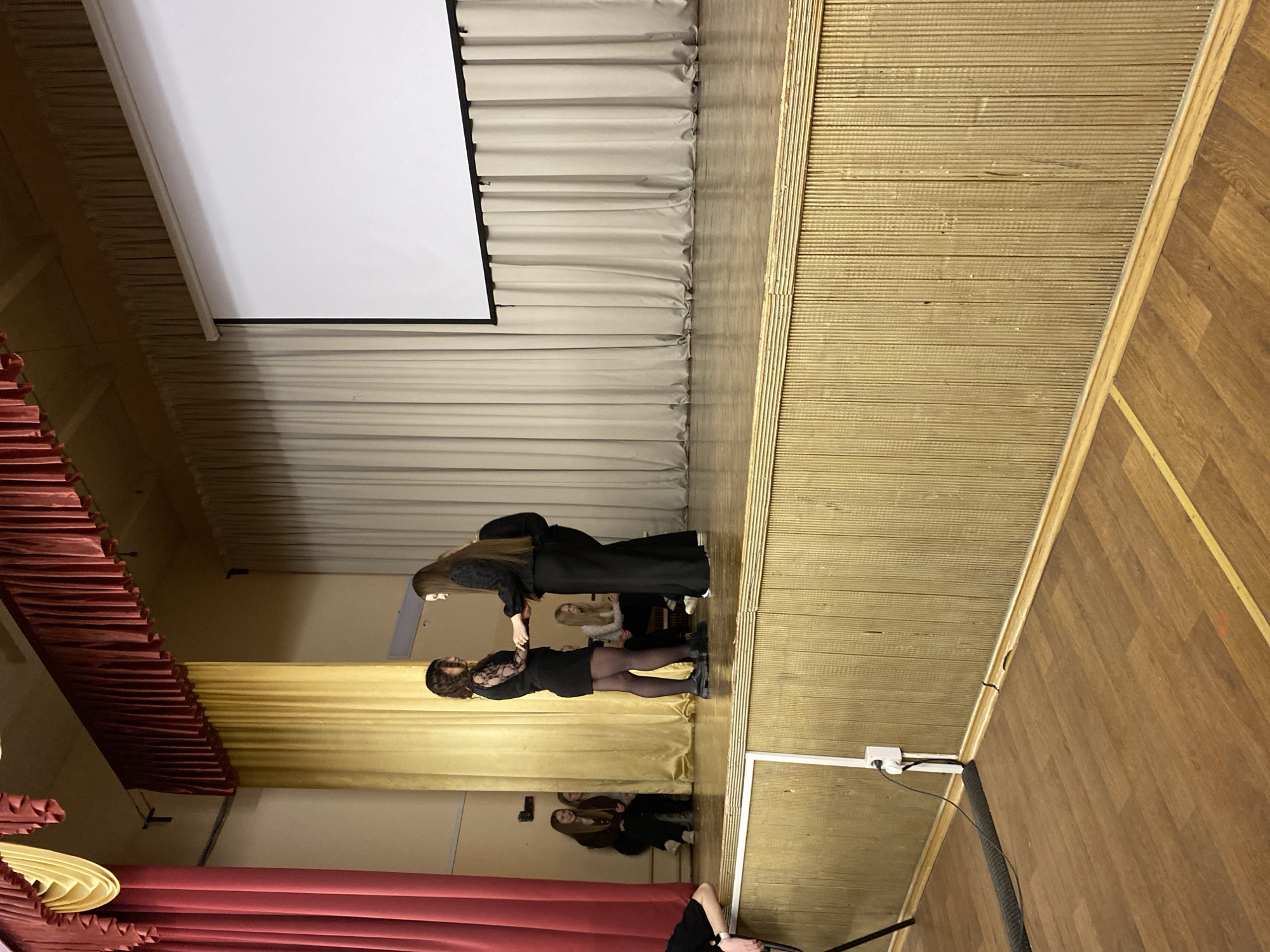 The image shows an indoor scene in what appears to be a stage or auditorium setting in Votkinsk, Russia. Two women in black clothing are the main subjects; one woman is extending her hand to assist the other. Behind them is a yellow curtain, and behind that, a group of seated individuals. Above, there is a white projection screen and more curtains. The stage has wooden flooring. Red curtains are visible towards the bottom of the frame. Electrical outlets are attached to the wall.