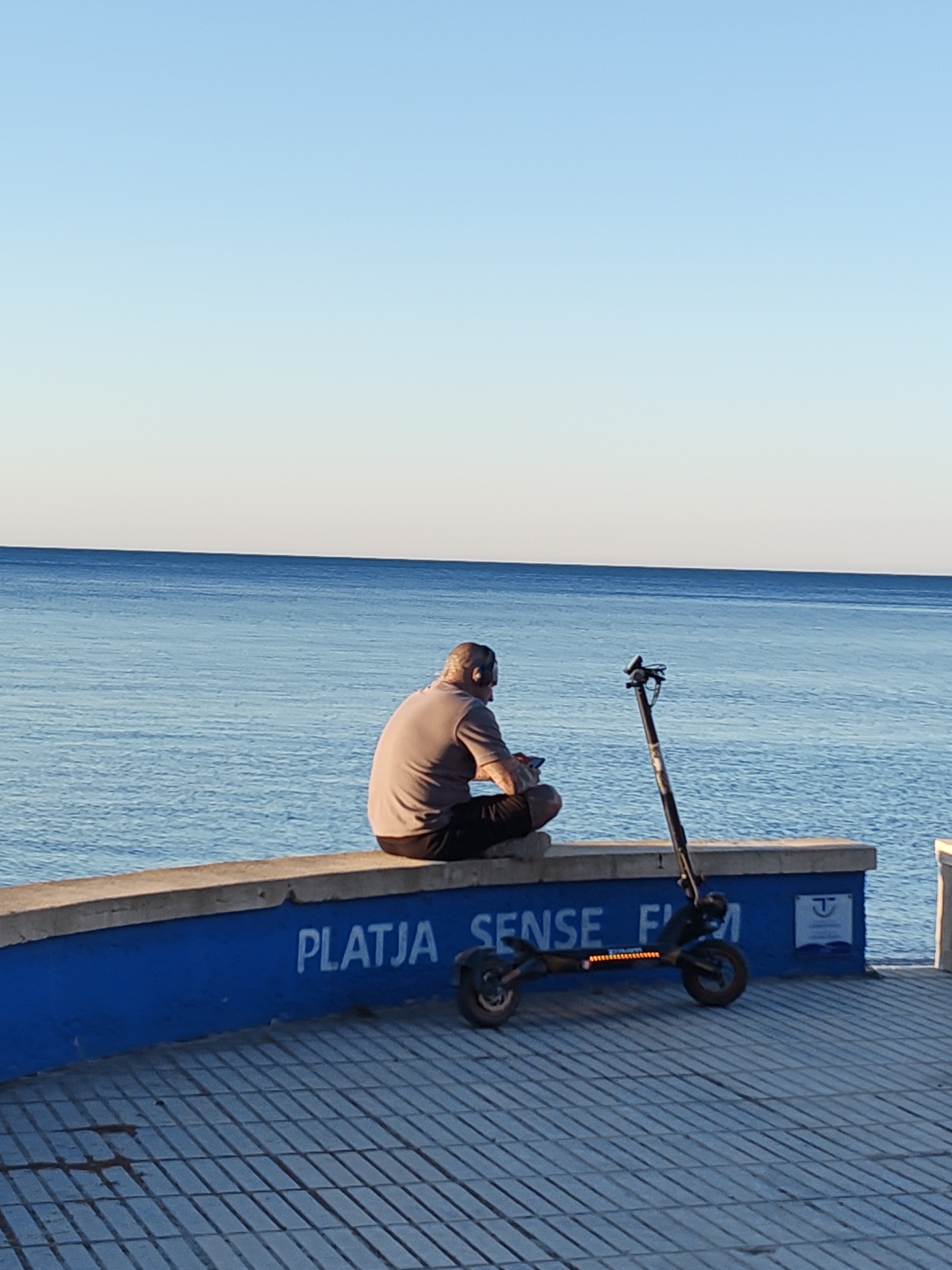 The image depicts a peaceful coastal scene, likely during late afternoon or early morning due to the soft, clear lighting. A man, seen from behind, is seated on a low concrete wall overlooking a tranquil blue sea. He wears a light-colored short-sleeved shirt and dark shorts, with headphones on his ears, and appears to be holding a small electronic device. His graying hair is visible.
Beside him, an electric scooter, black with visible orange lights along its side, is parked on a textured, patterned paved walkway. The concrete wall below the seating ledge is painted blue and features white text that clearly reads "PLATJA SENSE FUM," translating to "Smoke-free Beach" in Catalan/Valencian.
The background consists of a vast, calm blue sea meeting a clear, gradient sky at a distinct horizon line. The weather is clear and cloudless. The scene conveys a sense of quiet contemplation or relaxation by the water, possibly in Dénia, Spain.