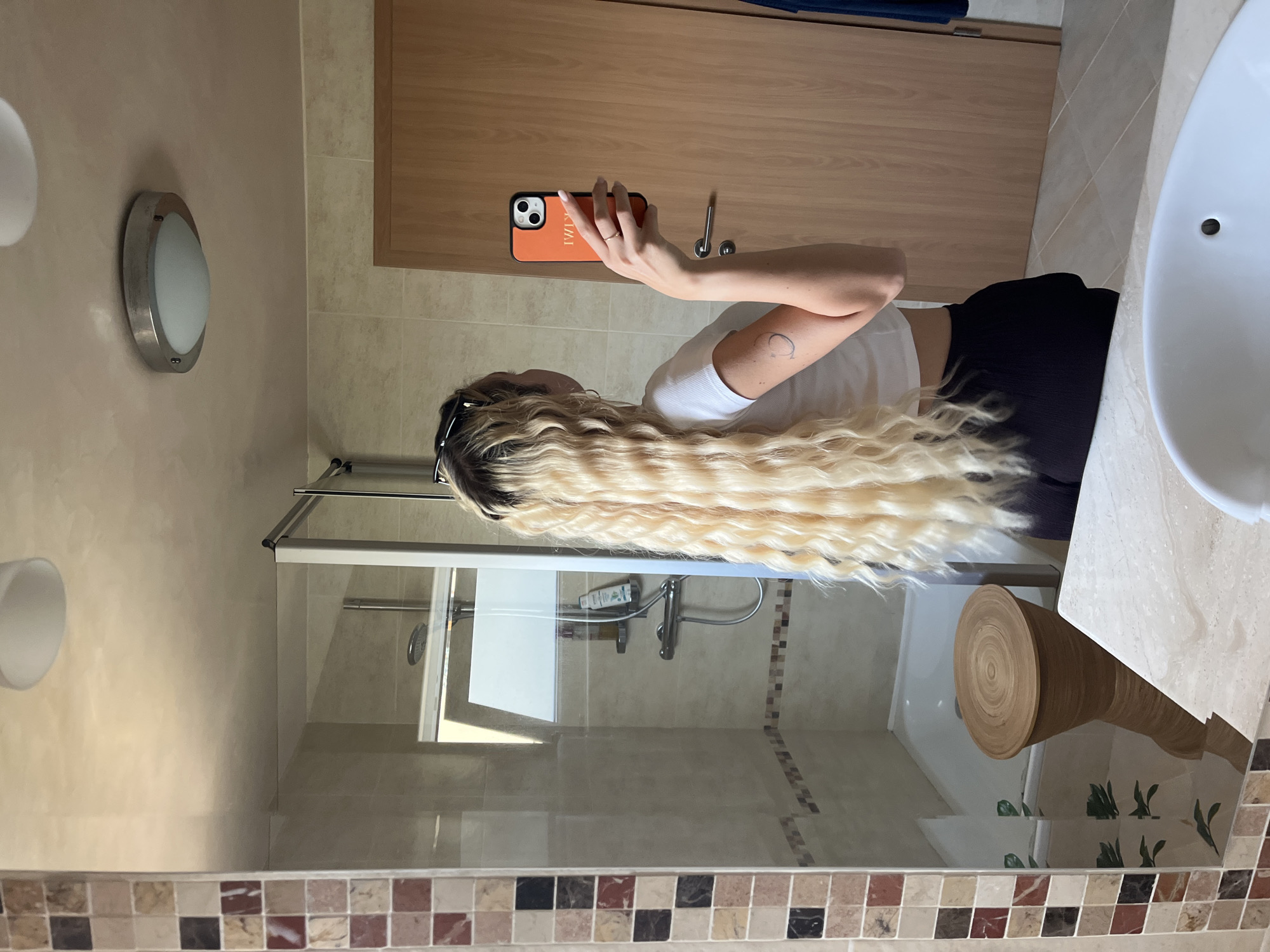 A mirror selfie is captured in a well-lit bathroom, showing a person from behind/side with very long, wavy, blonde hair that has visible dark roots. The person is wearing a white short-sleeved top and dark bottoms (shorts or trousers). Their left arm is raised, holding an orange-colored smartphone with a "KIMI" inscription written vertically in gold letters on its case. A small, circular tattoo is visible on their left upper arm.
The bathroom features light-colored wall tiles, a shower enclosure with a glass door and metal frame, and a white sink partially visible on the right. A silver-toned, oval-shaped wall light with a frosted cover is mounted on the wall to the left. In the reflection of the mirror, a wooden cabinet or door is seen behind the person. Near the floor, there's a mosaic tile border composed of various shades of brown, beige, and reddish tiles. A cylindrical wooden stool or decorative object with a visible spiral grain pattern on its top surface is positioned on the floor, and small green, leaf-like motifs or stickers are on the wall near the floor in the reflection. The overall setting suggests a modern indoor bathroom, likely in a residential home or hotel, illuminated by what appears to be bright daytime light combined with indoor fixtures.