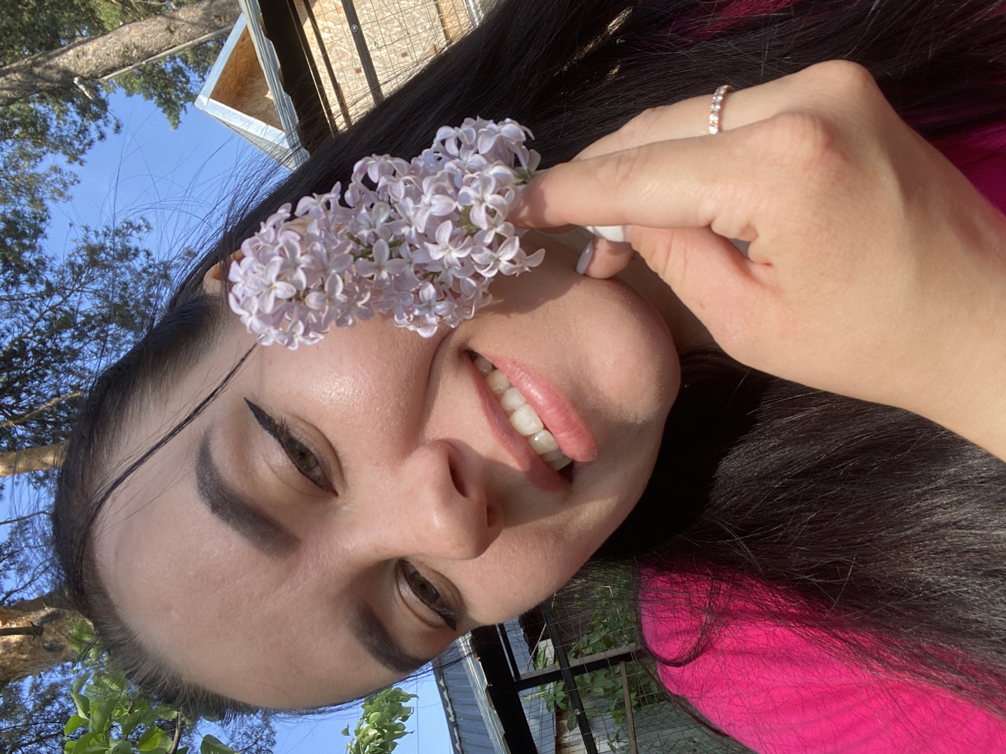 A woman poses in Chaikovsky, Russia, outdoors on a sunny day, holding a sprig of lilac flowers up to her left eye. She is wearing a bright pink shirt and a diamond ring on her right hand. Behind her are trees, the sky, a wooden shed, and a fence.