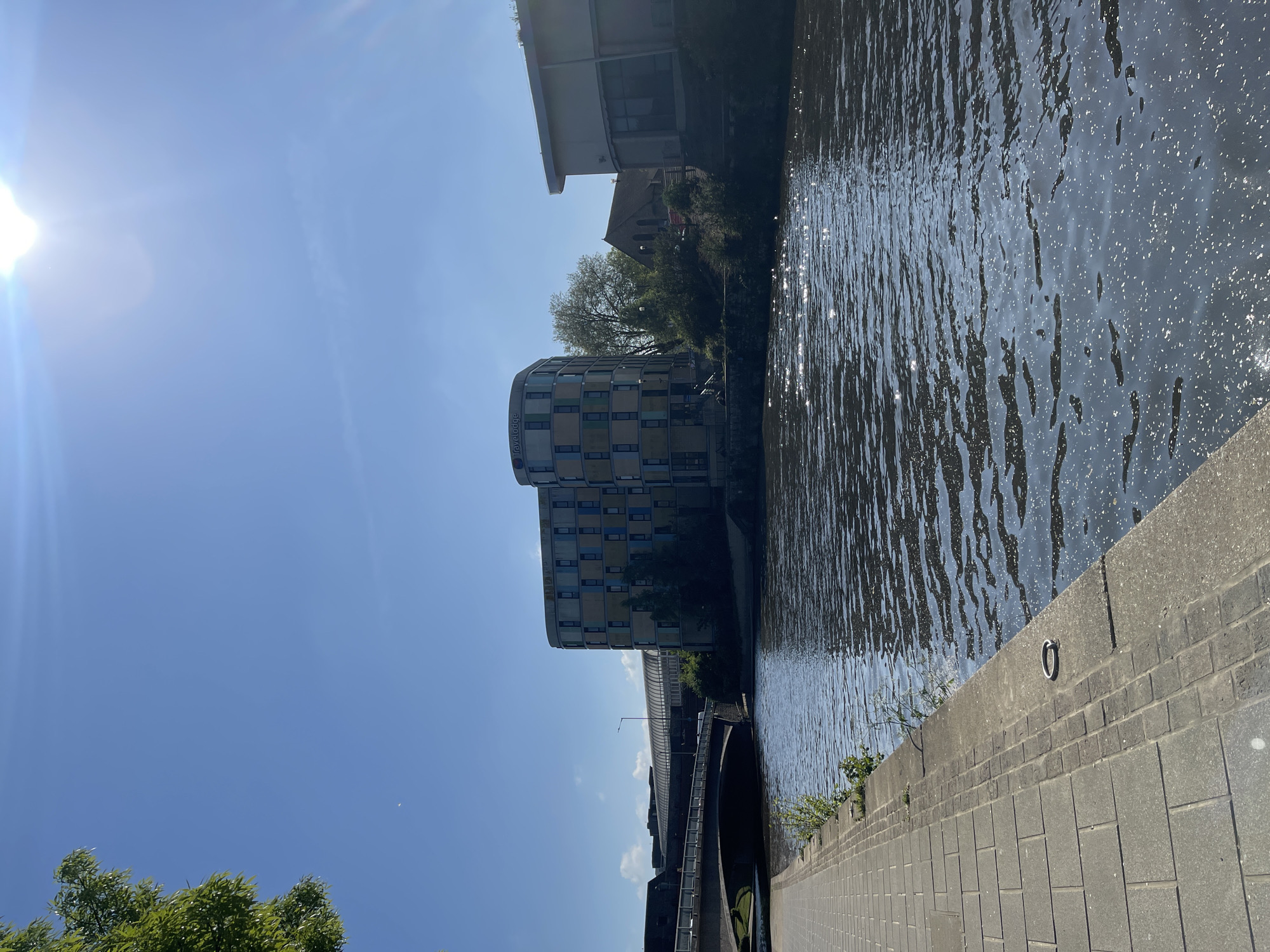 This media file captures an urban waterside scene in Maidstone, United Kingdom, under bright sunlight. The image is oriented vertically, with a body of water, likely a river or canal, running along the right side of the frame. The water surface shows numerous ripples and sparkling reflections from the sun.
To the immediate right of the water, a paved walkway made of grey rectangular blocks extends from the bottom to the top of the image. A concrete embankment wall separates the walkway from the water, featuring a visible metal mooring ring attached to it. Small patches of green vegetation grow near the base of this wall.
Across the water, several buildings and structures define the urban landscape. A prominent, modern, curved building stands out, characterized by multiple windows, some appearing to be balconies, and a facade made of blue, grey, and lighter-colored panels. The word "Travelodge" is clearly visible on the side of this building. Further back and slightly to the right of the curved building, a traditional structure with a pitched roof, possibly a church, is partially obscured by trees. Above the curved building, a long, horizontal structure, which could be a bridge or part of transport infrastructure, stretches across the middle ground.
The upper-left portion of the image is dominated by a clear, bright blue sky, where the sun is visible, causing a strong glare and lens flare. Some green treetops are visible in the bottom-left corner.
The scene is devoid of people. The strong sunlight and clear sky indicate a bright, sunny day. The image captures a static, calm moment of urban life by the water.