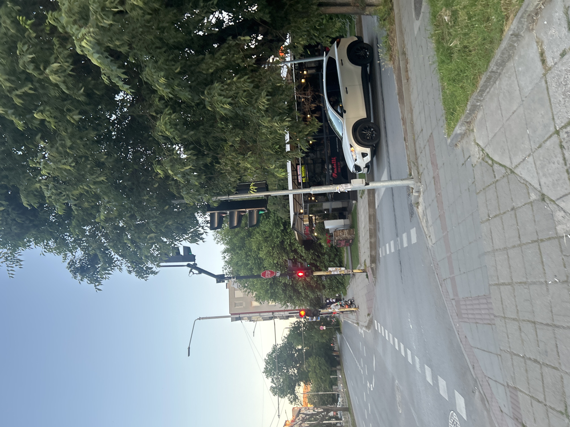 The image shows a street scene in Varna, Bulgaria. The scene appears to be an intersection, with traffic lights and crosswalks visible. A beige SUV is driving through the intersection. A person on a bicycle is also visible. The area is lined with trees and buildings. The time of day appears to be late afternoon or early evening, as the sun is low in the sky and casting long shadows.