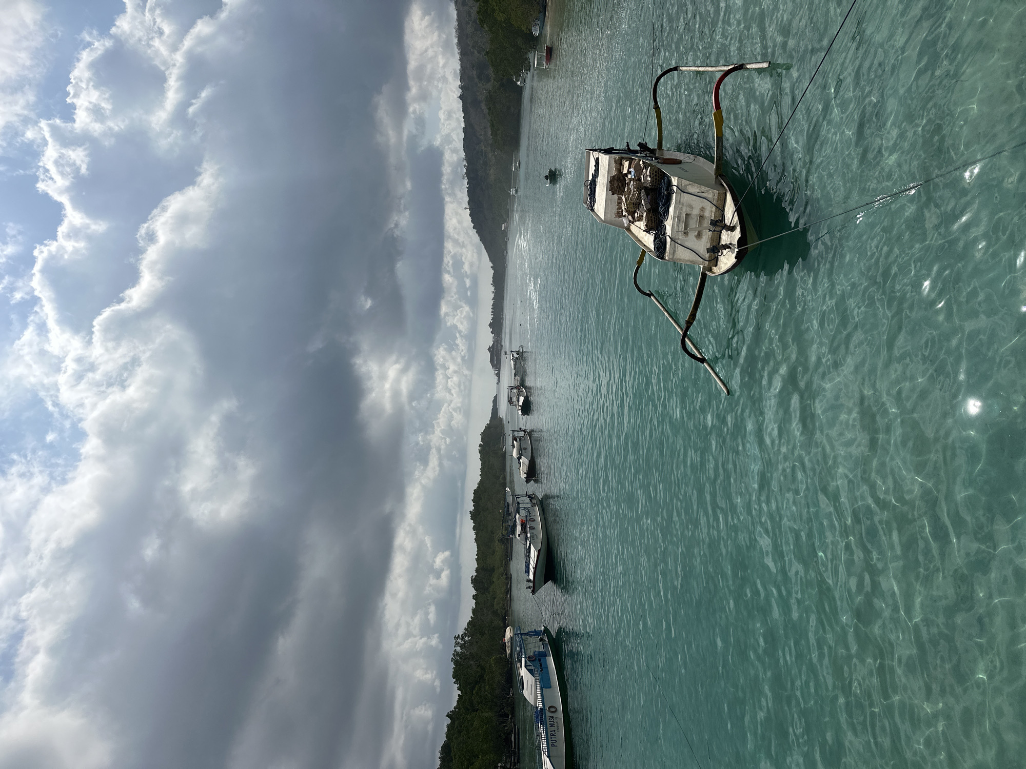This is a photograph taken in Nusa Penida, Indonesia. The image depicts a body of water, likely the ocean, with several boats moored or anchored. The water is a clear, turquoise color, with sunlight glinting off its surface. In the foreground, a smaller, traditional outrigger boat is prominently featured, loaded with baskets and coiled ropes. Further in the distance, a line of larger white boats, possibly passenger ferries or tour boats, are visible. The background shows a lush, green coastline with trees and hills. The sky is partly cloudy, with dramatic, dark storm clouds mixed with patches of blue sky and bright sunlight. The overall impression is a tropical coastal scene, possibly a harbor or a popular tourist spot, with varying weather conditions.