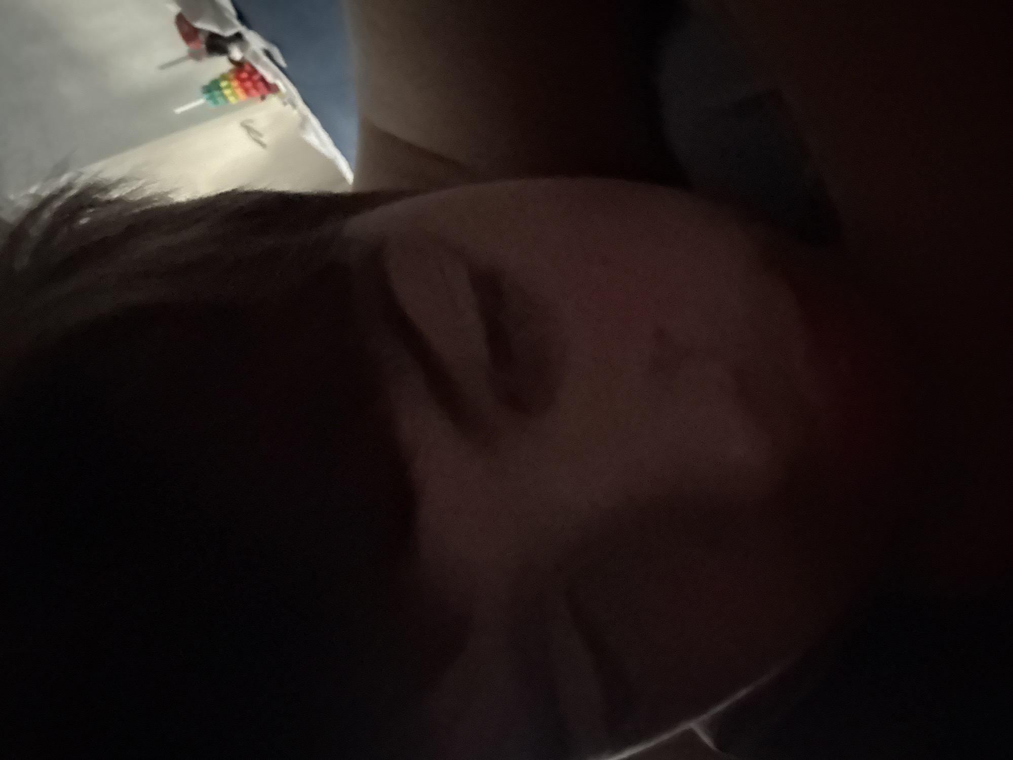 A close-up shot of a person's face in low lighting, seemingly resting or sleeping. The face is the primary subject, with features partially obscured by shadow. Some bedsheets and a toy-like object with a rainbow-colored component are visible in the background. The image suggests an indoor environment, possibly a bedroom. There is no information provided in the image to determine the location as the city of Unknown, Unknown.