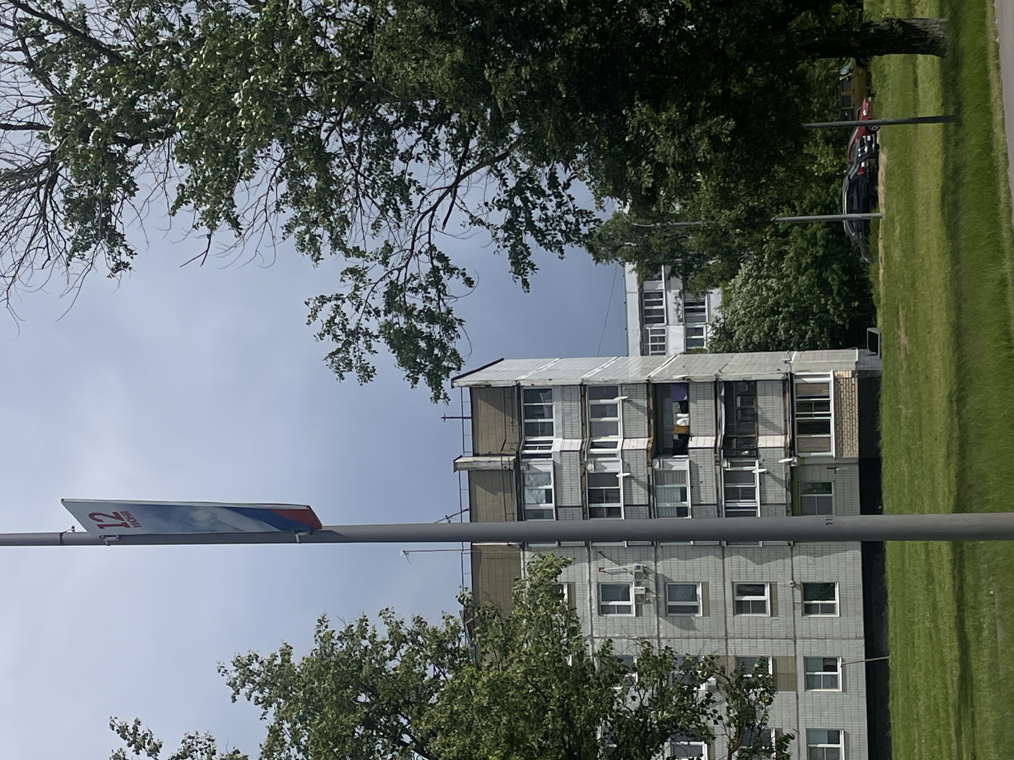 The image depicts a residential scene in Yakovlevskoye, Russia. The primary subjects are buildings and trees, suggesting an urban or suburban environment. A tall, multi-story apartment building, characterized by its gray, tiled facade and numerous windows, dominates the central portion of the frame. Balconies are visible on some floors. To the right of the building, a well-maintained, vibrant green lawn stretches out, indicating it is likely daytime and fair weather, possibly summer or late spring due to the lushness.
In the foreground and framing the scene are mature trees with dense green foliage, suggesting a warm season. A street sign, which appears to be a bus route marker, is attached to a pole crossing the image horizontally. The sign is rectangular and white, with blue and red stripes along its border. Text on the sign is visible but partially obscured, though the numeral "12" is clearly legible.
Behind the apartment building, more trees and other structures are visible, including another apartment building further in the distance. Several cars are parked on the grass or along the edge of a road, partially obscured by trees and foliage, indicating a residential area where parking is available. The overall impression is one of everyday life in a Russian residential district. The lighting suggests it is daytime, with a bright sky and natural light illuminating the scene. No people are directly visible, and the scene is peaceful and static.