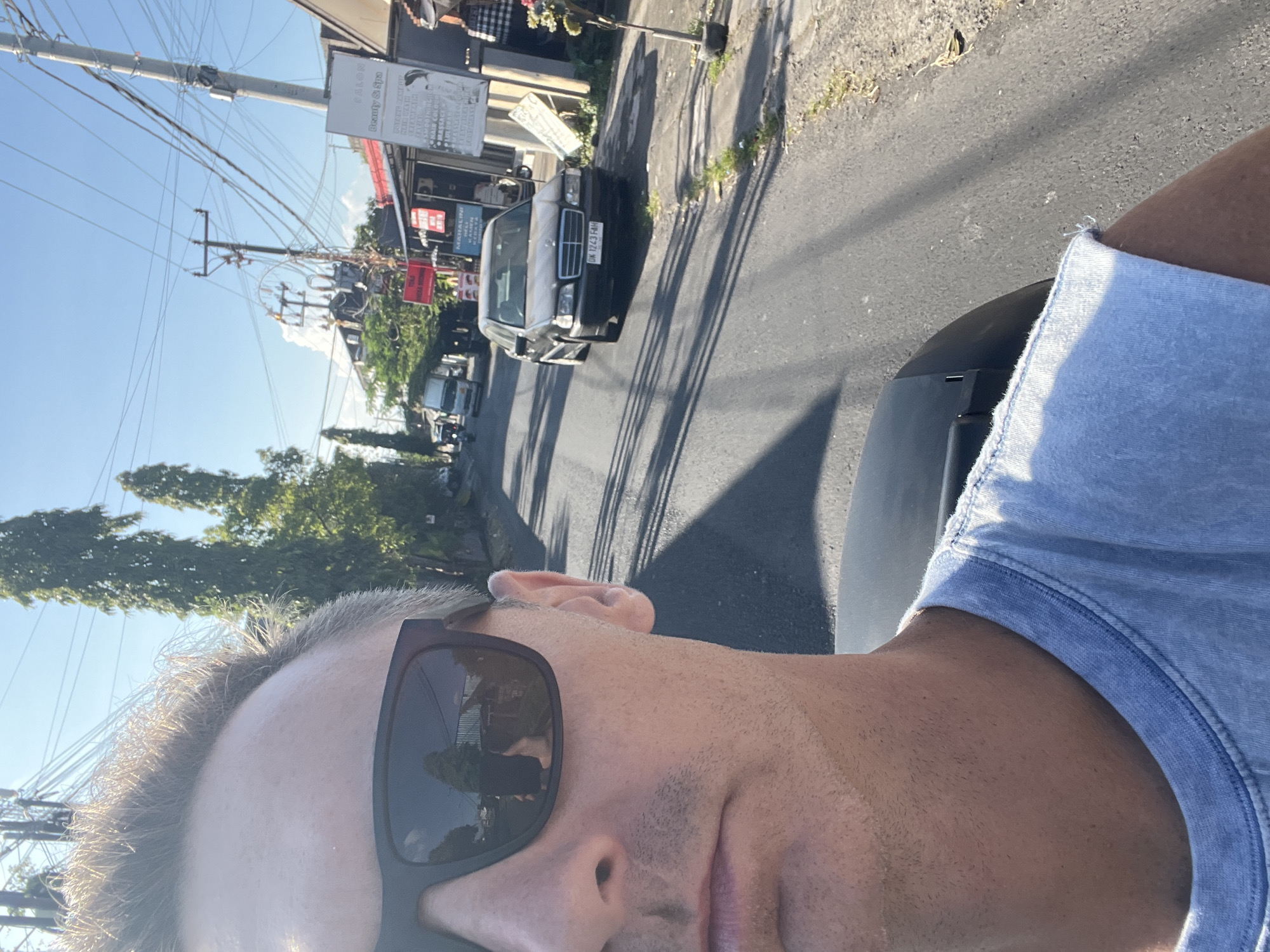 A man wearing sunglasses and a light blue t-shirt is taking a selfie in a street in Sukawati, Indonesia. He is in the foreground, with the road and buildings on either side extending into the background. There is a dark-colored car with license plate DK 1243 FAH parked on the right side of the road, and a truck is visible further down the street. Trees and power lines line the street, and the sky is clear and blue. The scene appears to be a typical street view in a tropical urban environment.