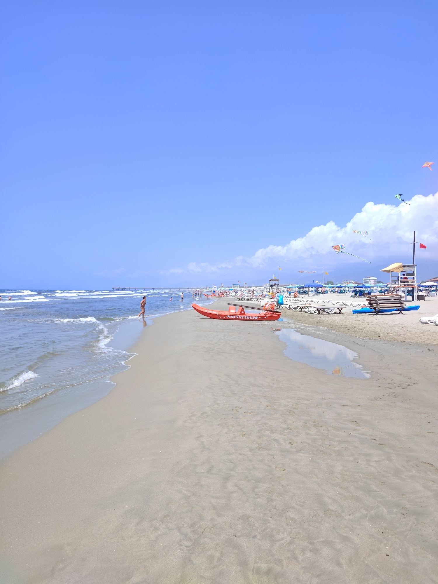 The image depicts a crowded beach in Pietrasanta, Italy, on a bright, sunny day with a clear blue sky. The foreground shows a sandy beach with gentle waves lapping at the shore. In the middle ground, there are several beach chairs and umbrellas, suggesting a popular tourist spot. A bright red life raft with the word "SALVAGGIO" written on it is visible on the sand, indicating a potential safety presence. In the background, a pier extends into the sea, and more people can be seen enjoying the beach. Kites are flying in the sky, adding a festive touch to the scene. The overall impression is one of a lively and enjoyable summer day at the beach.