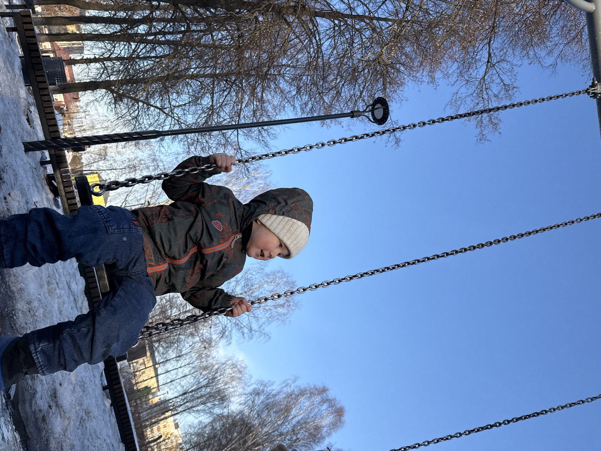 A child is on a swing in what appears to be a playground in Zav'yalovo, Russia. The child is wearing a winter coat and hat, and the ground has patches of snow. The sky is blue. Bare trees are visible in the background. A bench is near the swing set.
