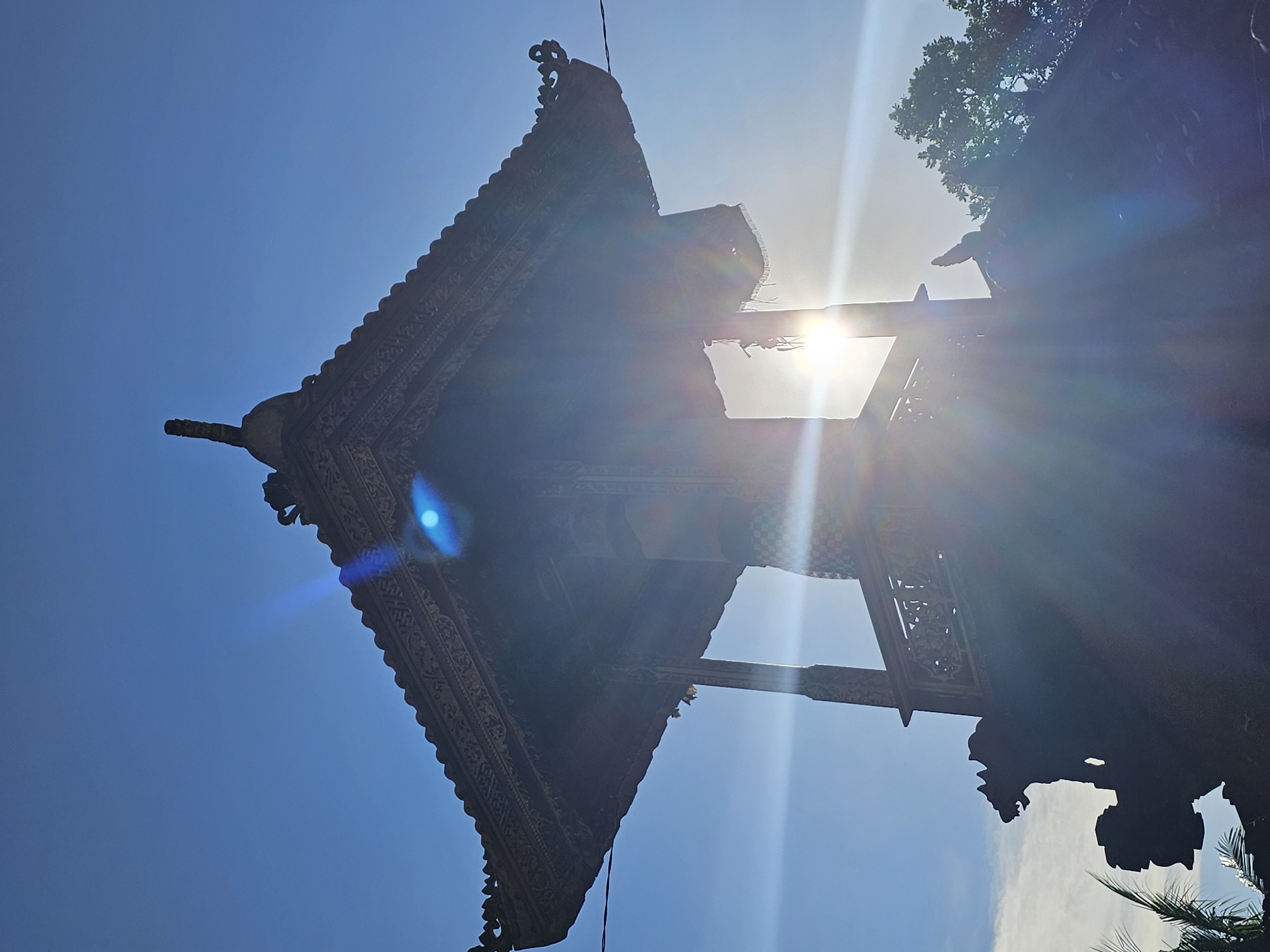 Looking up at a Balinese structure in Kuta Selatan, Indonesia, the image captures a building with intricate carvings and a distinctive roofline silhouetted against a bright, blue sky. The sun is positioned directly behind the building, creating a strong glare that obscures some details. The building's architectural elements are visible, indicating traditional Balinese craftsmanship. Parts of the environment, including trees, are also visible.