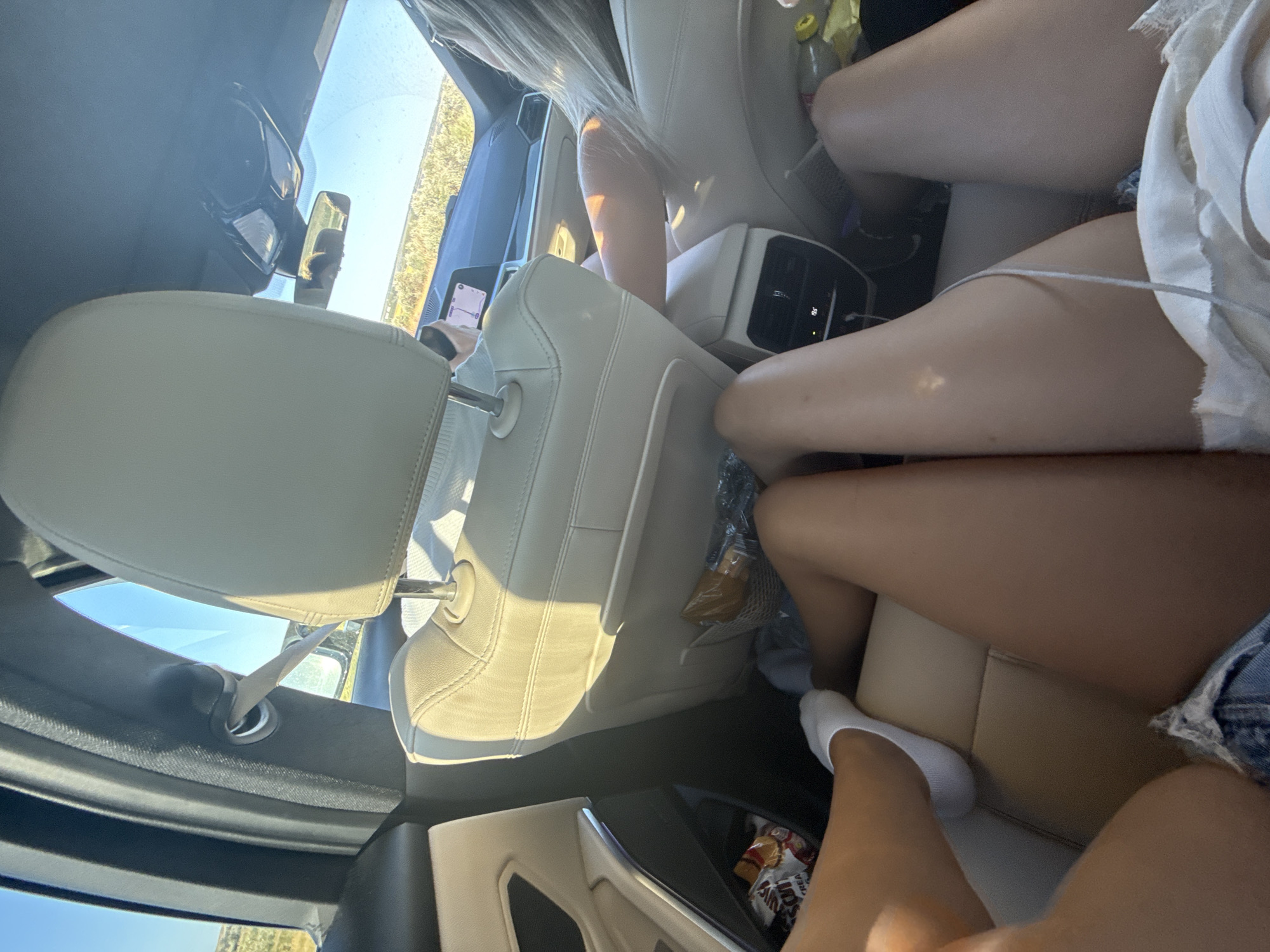 In an unknown city, a shot captures the interior of a car during the day. The perspective is from the backseat, focusing on the legs of two individuals. One person is wearing white socks and denim shorts. The other person's legs are also visible. The car's interior is adorned with beige leather seats and a black dashboard. Snacks are placed in the seat pocket. The sunlight streams in through the windows, illuminating the scene.