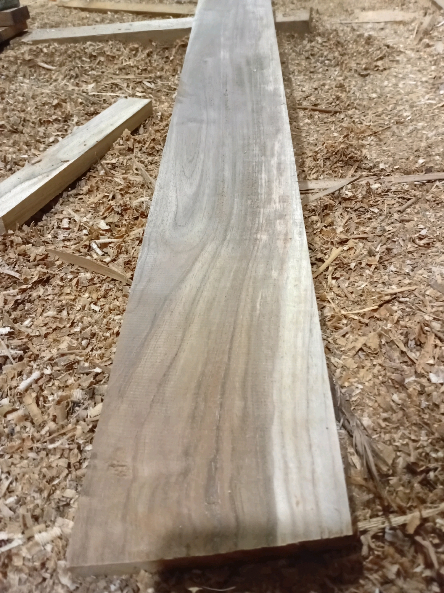 A close-up, eye-level shot shows a long, narrow wooden plank lying horizontally on a bed of wood shavings. The plank is smooth and light brown with visible grain patterns. To the left of the main plank, a shorter, thicker piece of wood is angled, also resting on the shavings. The background is filled with a dense layer of light brown wood shavings, with hints of other wooden objects and surfaces visible further back, suggesting a woodworking or lumberyard environment. The scene is devoid of people or any visible activity. The location is identified as Playen, Indonesia.