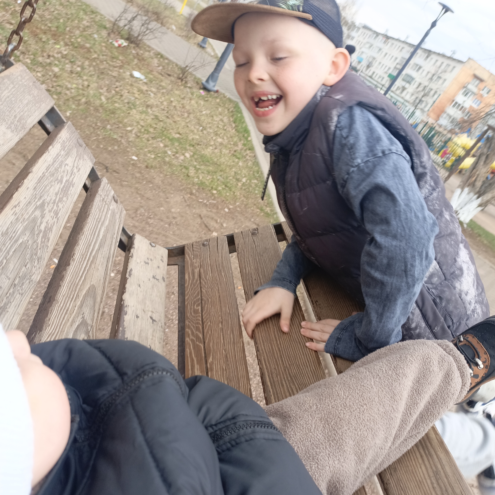The image depicts a child wearing a baseball cap, a vest, and a blue shirt, laughing and leaning against a wooden bench. Another child, primarily visible by their head and upper body, is also seated on the bench wearing a black jacket. The setting appears to be a playground or park area in an urban environment. Buildings are visible in the background, and there is foliage and playground equipment in the surrounding area.