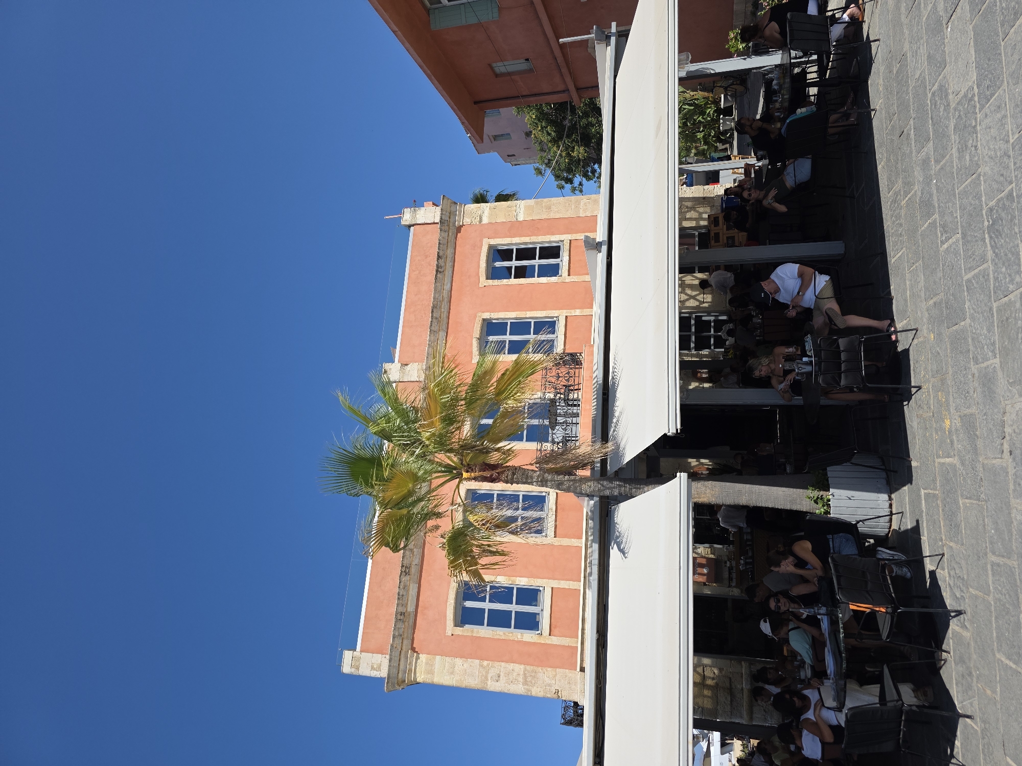 In the city of Chania, Greece, a sun-drenched outdoor cafe scene unfolds. The main subject is a two-story, salmon-colored building with cream-colored stone accents, featuring large blue-paned windows and decorative balconies. A tall palm tree with vibrant green fronds stands in front of the building, partially obscuring the upper floor.
The ground floor of the building is occupied by a cafe with white retractable awnings providing shade to outdoor seating. Numerous patrons are seated at small, dark-colored tables, some of which have umbrellas. People are engaged in various activities: conversing, looking at their phones, and appearing to enjoy their meals and drinks. The cafe's interior is visible, showing a bar area with staff attending to customers.
The cafe seating area extends onto a paved patio made of gray cobblestones. To the right of the main building, another similarly colored building is visible, also with windows and architectural details. The sky above is a clear, bright blue, indicating a sunny day. The overall impression is a lively, relaxed atmosphere typical of a European seaside town.