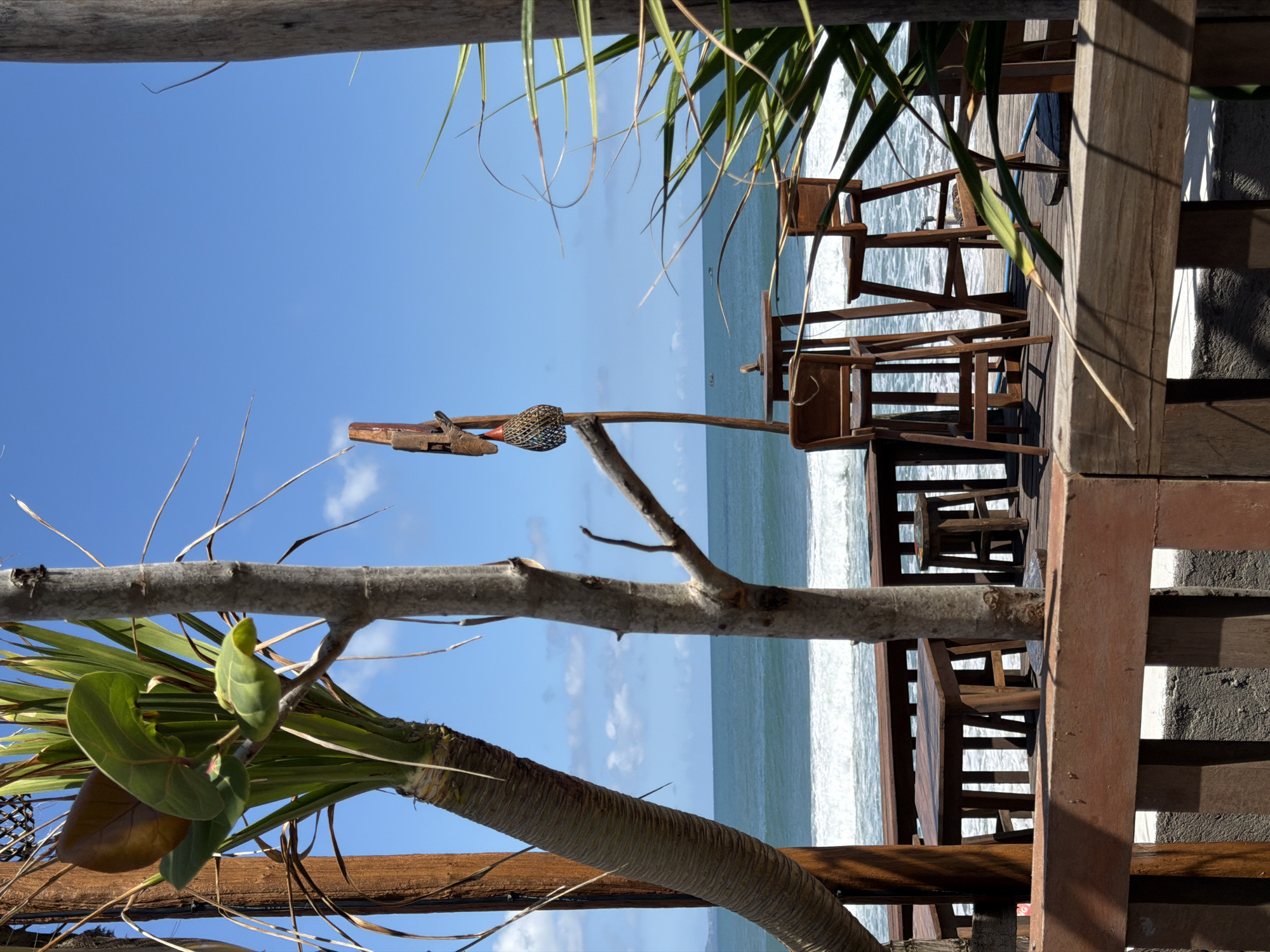 The scene depicts a view from a wooden structure, possibly a restaurant or deck, overlooking the ocean. The sky is bright blue with some clouds. The ocean has waves. The view is partially framed by a tree with green and brown leaves and a textured trunk. There are wooden chairs and structures on the deck, suggesting an outdoor seating area. In North Kuta, Indonesia.