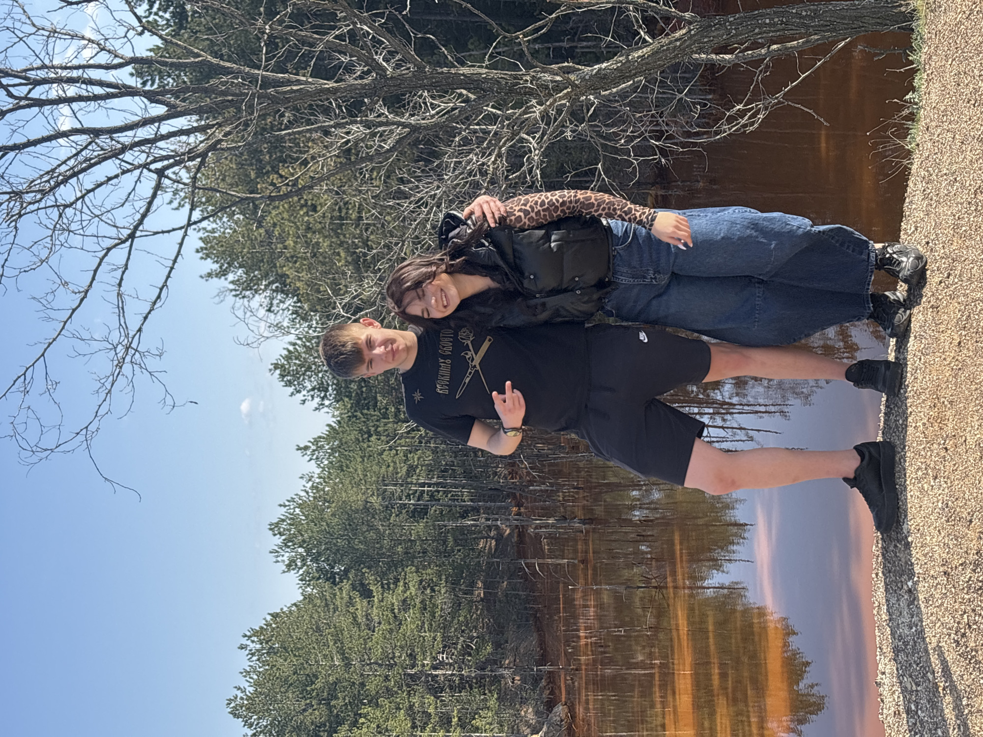 Two individuals are posing in front of a body of water and a forest landscape. A young man is wearing a black t-shirt and black shorts, while a young woman is wearing a leopard print long-sleeved shirt and a black vest. The woman is leaning on the man's shoulder. The photo was taken in Elshitsa, Bulgaria.
