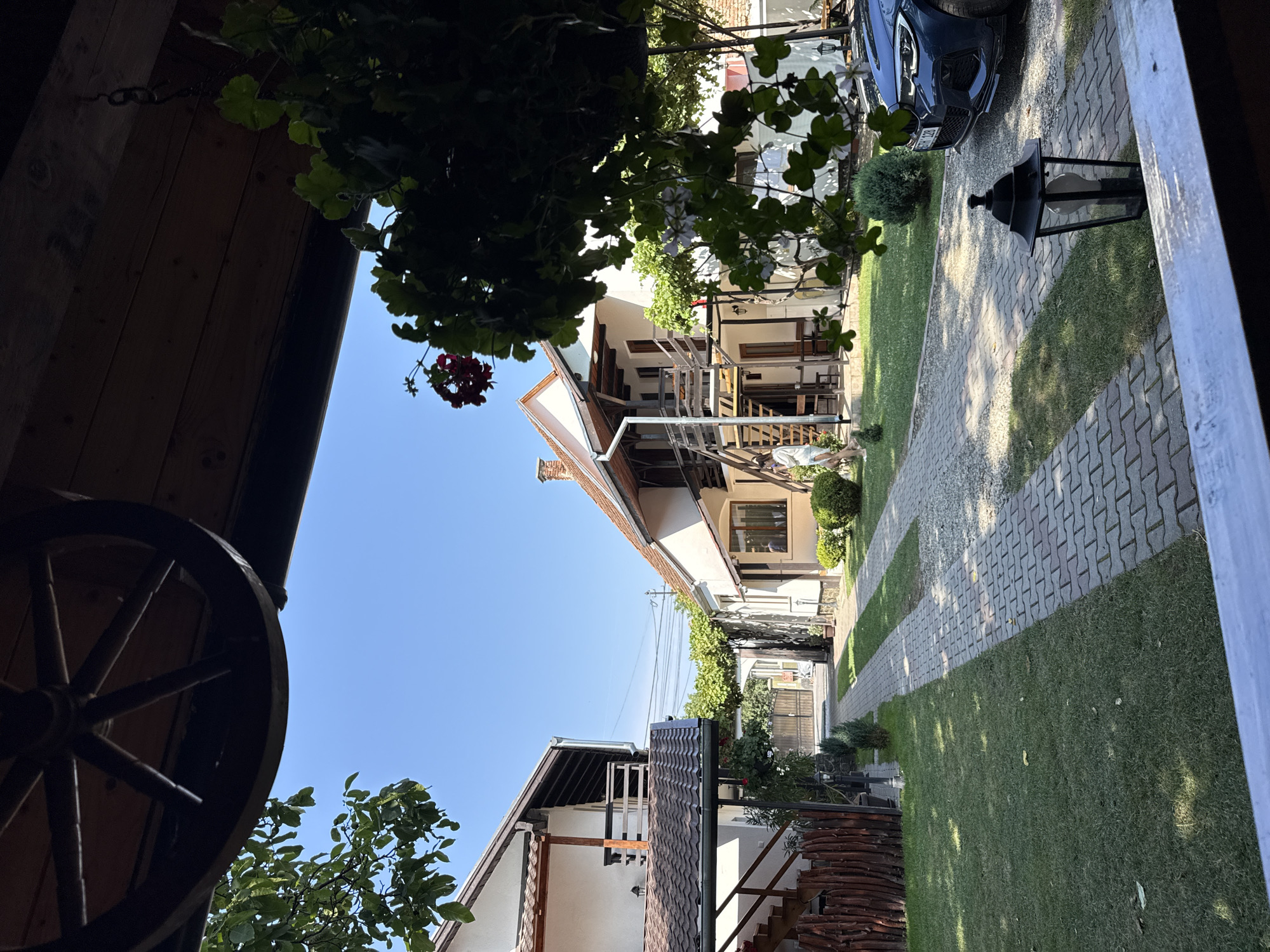 The image depicts a courtyard scene in Cârțișoara, Romania, likely a guesthouse or private residence. Two white buildings with reddish-brown tiled roofs flank a paved driveway that extends towards a wooden gate in the distance. The building on the left features a wooden staircase leading to an upper-level balcony. The building on the right has a similar architectural style, with an external staircase and wooden balconies. Lush green grass covers the courtyard, with some scattered shrubs and trees. A dark blue car is parked on the right side of the driveway. A decorative wooden wagon wheel hangs from the overhang of a covered porch in the foreground, partially obscuring the view of the sky. Hanging plants with red and white flowers are also visible in the foreground, adding a touch of natural beauty to the scene. A person, appearing to be a woman wearing light-colored clothing, is walking on the grass near the building on the right. A traditional lantern sits on the paving stones near the foreground. The overall atmosphere suggests a serene and well-maintained outdoor space.