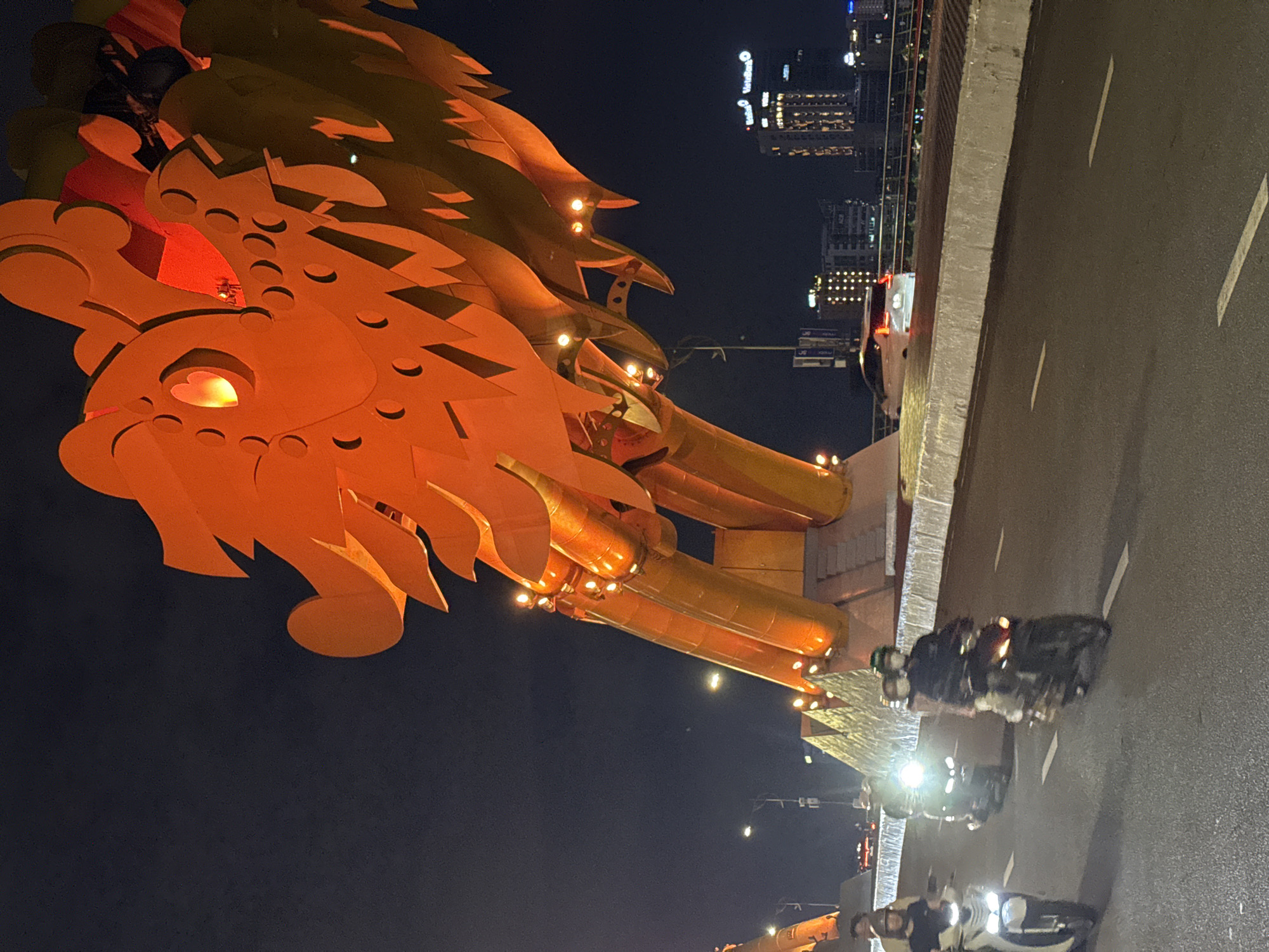 The image captures a nighttime scene in Da Nang, Vietnam, featuring the illuminated Dragon Bridge. The bridge's iconic dragon sculpture is prominently displayed, its orange and yellow scales glowing under the night sky. The dragon's head is a focal point, with its eye glowing warmly.
Several motorcycles are visible on the bridge's roadway, with riders and passengers illuminated by the streetlights and headlights. One motorcycle in the foreground has its headlights on, casting bright beams onto the wet asphalt. The road surface appears slightly reflective, suggesting recent rain or damp conditions.
In the background, the city skyline of Da Nang is visible, with numerous buildings lit up. The lights create a dazzling display against the dark sky. The overall atmosphere is lively and vibrant, characteristic of a city at night. The time of day is clearly night, and the location is identifiable as Da Nang, Vietnam, due to the presence of the Dragon Bridge. The mood is one of urban energy and the dynamism of city life at night. No discernible text is visible in the image.
