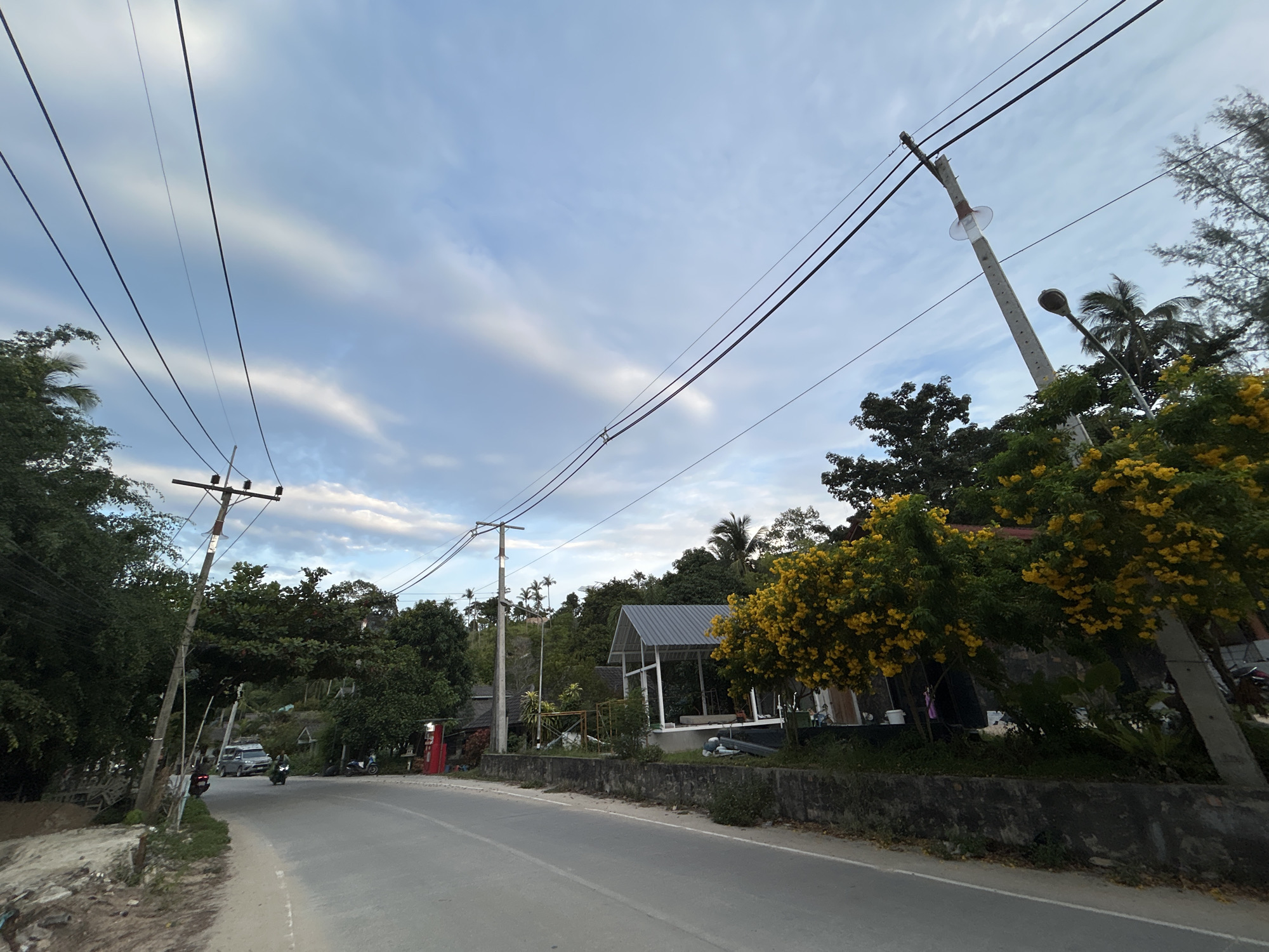 A paved road curves through a tropical landscape on Ko Pha-ngan, Thailand, under a sky filled with streaky clouds. Overhead, power lines stretch across the frame, supported by utility poles. Dense green foliage, including palm trees and trees laden with bright yellow flowers, lines the side of the road. In the background, a modern-looking open-air structure with a gray metal roof sits nestled among the trees. A few buildings, some with red accents, are visible further in the distance.
On the road, a silver van drives away from the viewer, followed by a person on a motorcycle. A parked motorcycle is also visible on the right side of the road. The scene appears to be set in the late afternoon or early evening, as indicated by the soft, diffused light and the cloud formations. The overall atmosphere is peaceful and rural, with a touch of tropical charm due to the abundant flowering trees. No visible text is present in the image.