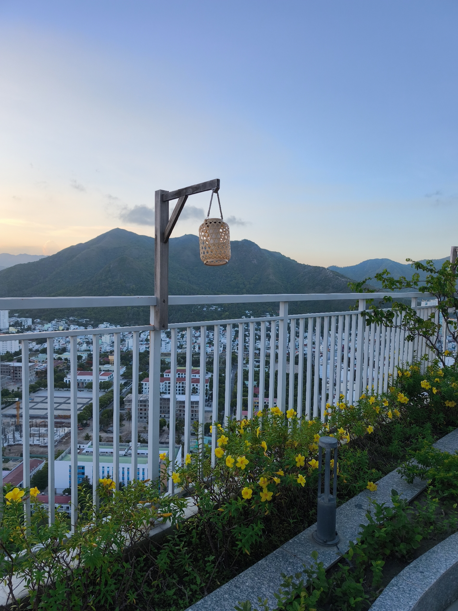 The image is a wide shot of a balcony or rooftop in Bac Nha Trang Ward, Vietnam, overlooking a city and a mountain range. The sky is a clear, pale blue with hints of sunset glow near the horizon. A wooden post with a woven bamboo lantern hanging from it stands in the foreground. A white railing runs along the edge of the balcony, with green foliage and yellow flowers growing in front of it. The city below is a dense collection of white and light-colored buildings, interspersed with darker green trees. In the distance, rolling green mountains form the backdrop. A modern-style grey light fixture is visible on the pathway to the right of the frame, with decorative stone borders. The scene appears tranquil and scenic, likely captured at dusk.