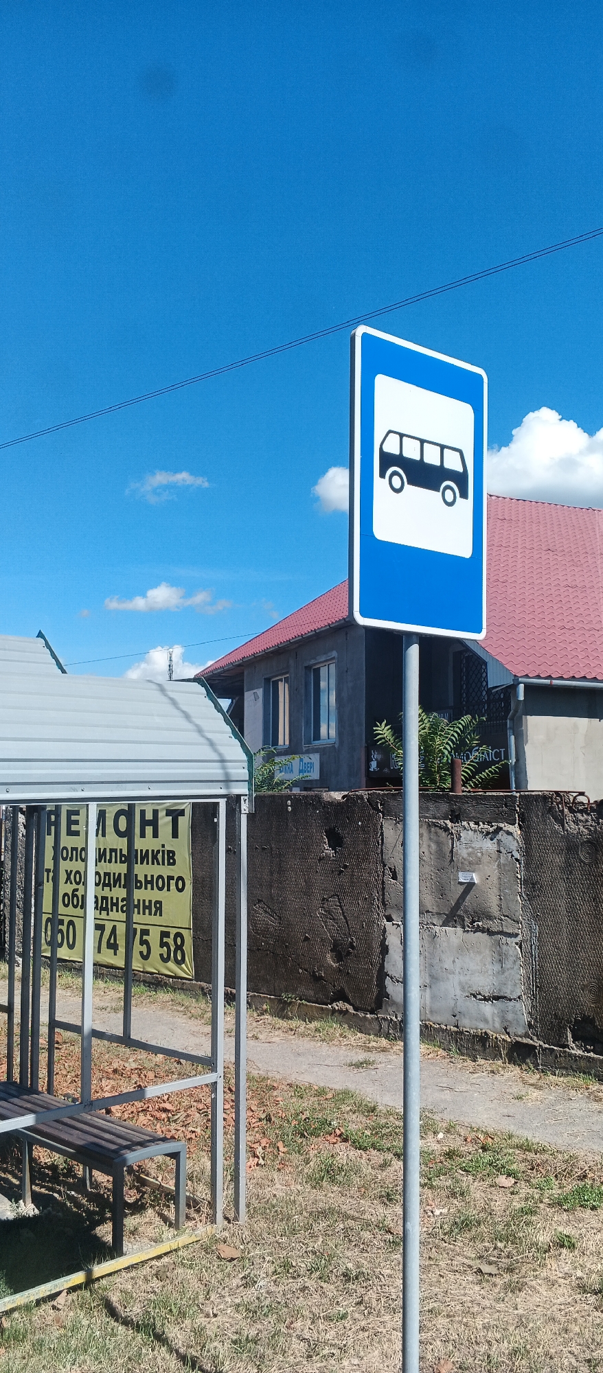 A bus stop sign is prominently displayed in the foreground, featuring a blue background with a white square containing a black silhouette of a bus. The sign is mounted on a gray metal pole. To the left of the sign, a bus shelter with a gray metal roof and bench is partially visible. A yellow sign with Ukrainian text and a phone number is attached to the shelter's frame. In the background, a gray building with a red roof stands behind a textured concrete wall. The environment is an outdoor scene with dry grass and scattered leaves on the ground, under a bright blue sky with some white clouds. The location is identified as Mukachevo, Ukraine.
