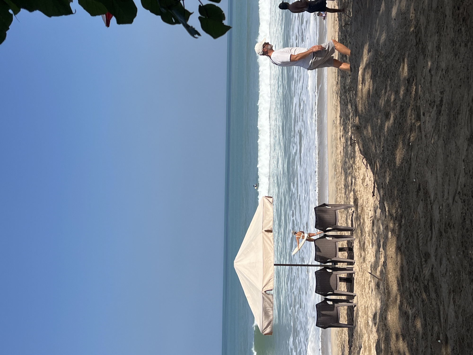 The image depicts a beach scene in Kuta, Indonesia. In the foreground, there are several empty plastic chairs arranged on the sandy beach, casting shadows from overhead foliage. Further back, a woman in a white bikini is standing by a large beige beach umbrella, holding a surfboard. She appears to be looking towards the ocean. In the mid-ground, waves are crashing on the shore. Two men are walking along the beach in the right mid-ground. One man, wearing a white t-shirt, grey shorts, and a white cap, is walking away from the viewer. Another man in dark shorts is walking further down the beach. The ocean stretches to the horizon under a clear blue sky.