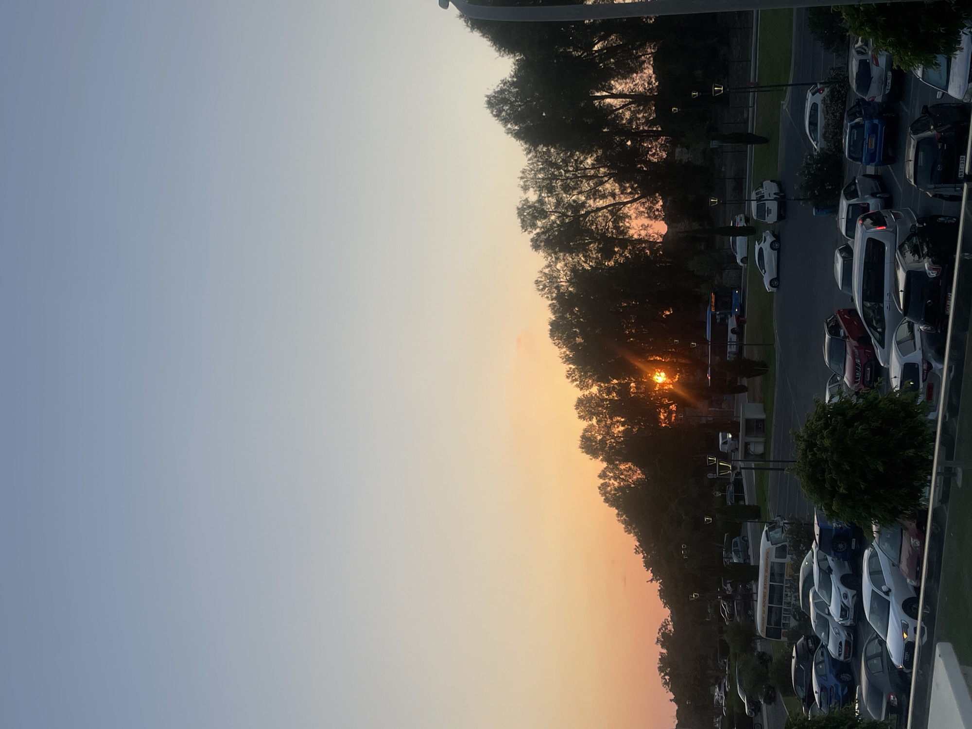 The image captures a sunset over a parking lot in Paphos Municipality, Cyprus. The sky transitions from a pale blue at the top to a warm orange and pink hue near the horizon, where the sun is setting behind a line of trees. The parking lot is filled with numerous cars, including sedans, SUVs, and a bus labeled "HERAKLES BATHS WATERFALLS." Streetlights are visible throughout the parking area. The overall scene suggests an outdoor public space in the late afternoon or early evening.