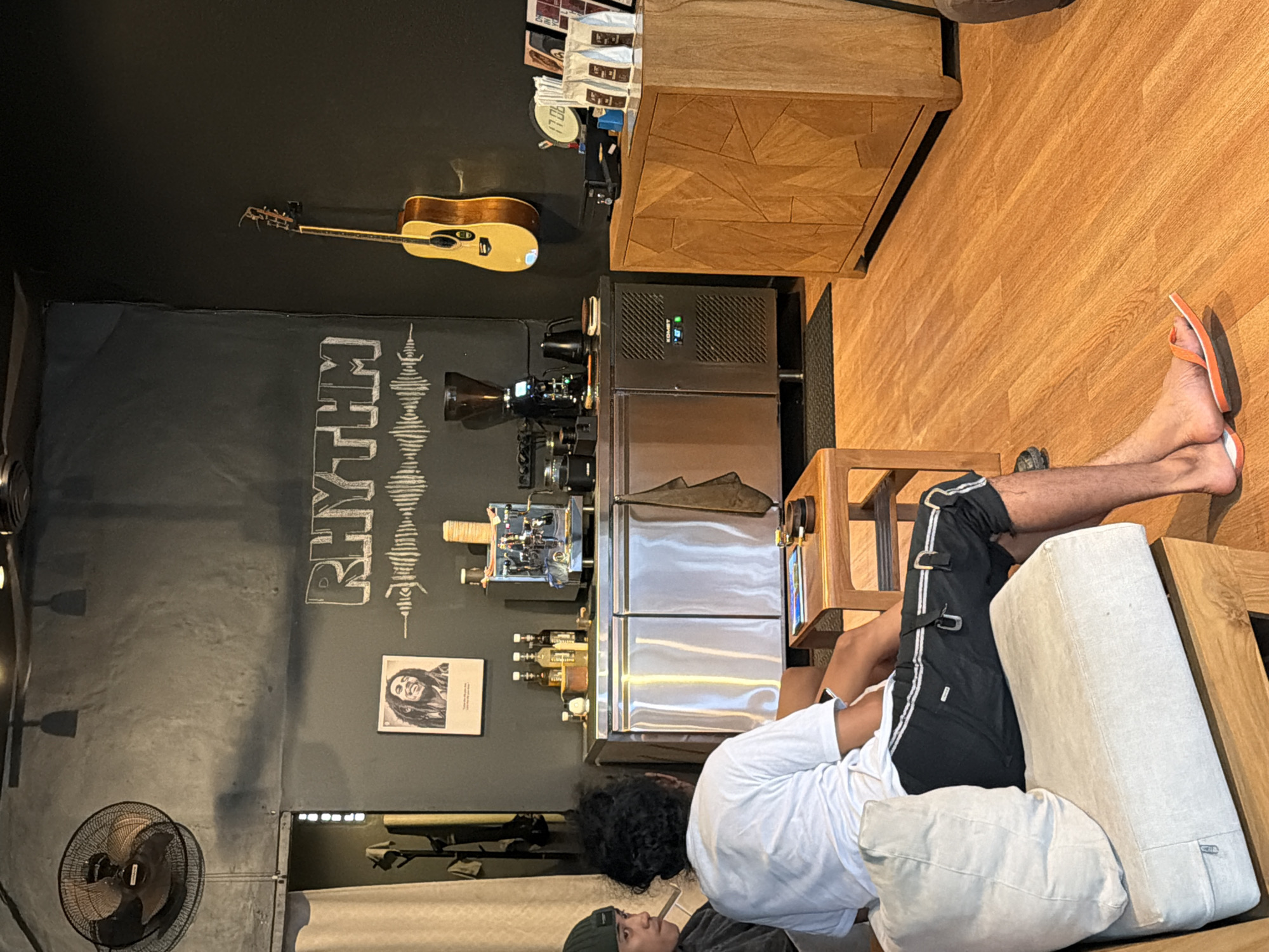 A sideways indoor shot shows a man reclining on a light-colored sofa in what appears to be a café in Kuta, Indonesia. The man wears a white t-shirt, dark shorts with white stripes, and orange flip-flops. He rests on light-colored cushions and has dark curly hair.
The café has dark walls with the word "RHYTHM" in a stylized font and graphic representations of sound waves. A guitar hangs above a coffee machine and associated equipment. A poster of Bob Marley is visible.
The floor is light-colored wood, and a wooden cabinet sits near the coffee machine. A black fan is in the lower-left corner. Another person wearing a dark green beanie and dark clothing is visible on the left side of the sofa.