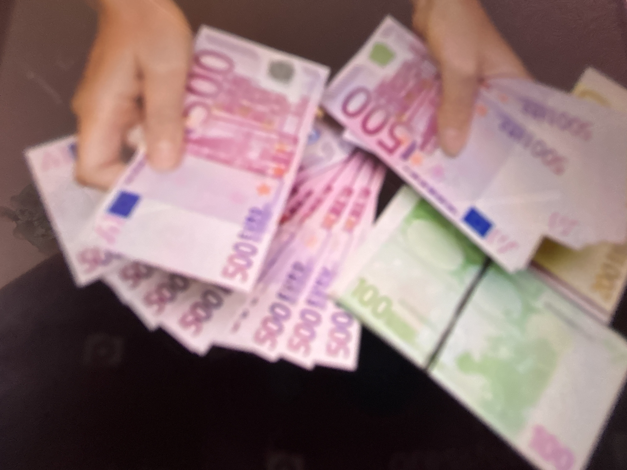 This media file shows a close-up, blurred image of hands holding and fanning out a large sum of Euro banknotes. The denominations visible include 500 Euro notes, indicated by the prominent "500" text on many of the bills. There are also some 100 Euro notes, identifiable by the green color and "100" text. The hands are lightly colored, suggesting fair skin. The background is dark and indistinct, offering no specific environmental cues. The lighting is somewhat dim and diffused, contributing to the overall blurriness. The image appears to be taken indoors, with no indication of the time of day or weather conditions. The activity depicted is the handling of a significant amount of cash, which could imply a transaction, a display of wealth, or simply someone counting money. There is no identifiable emotion conveyed due to the blur. The visible text consists primarily of the numerical denominations of the Euro currency. Given the provided location context of Gelendzhik, Russia, the presence of Euros suggests international currency or potential economic activity related to tourism or trade in that region.